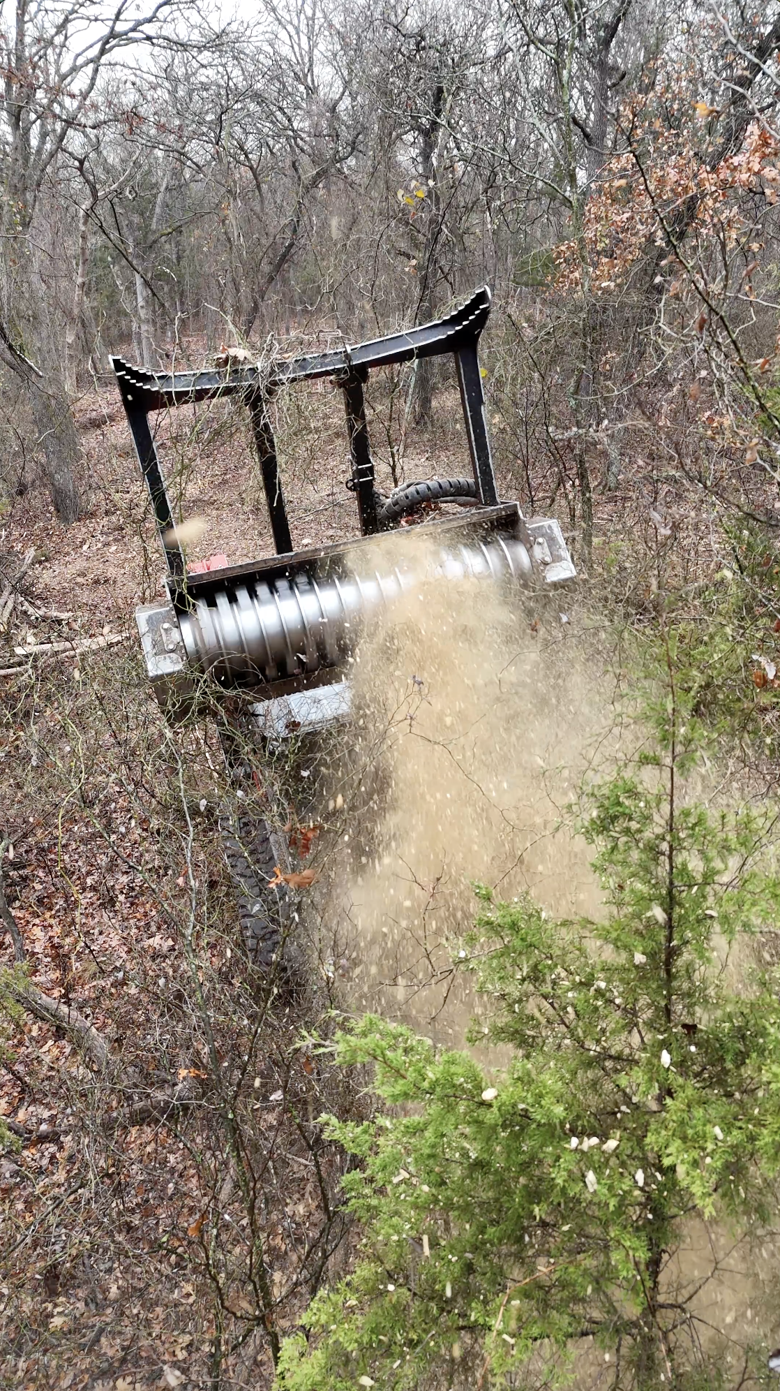 A large mechanical brush cutter clearing or trimming a dense area of bushes and small trees in a wooded area during daytime.