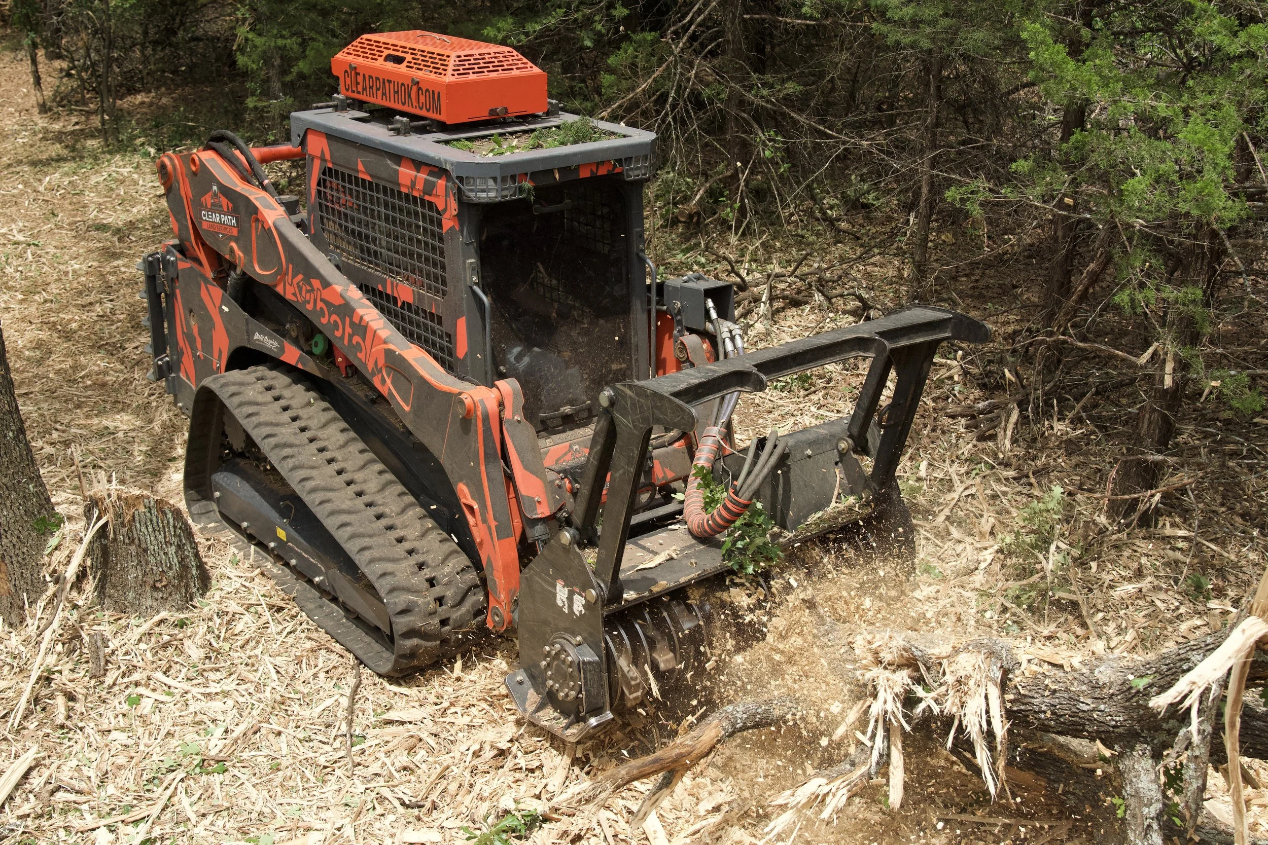 A small orange and black tracked machine clearing trees and debris in a forested area.
