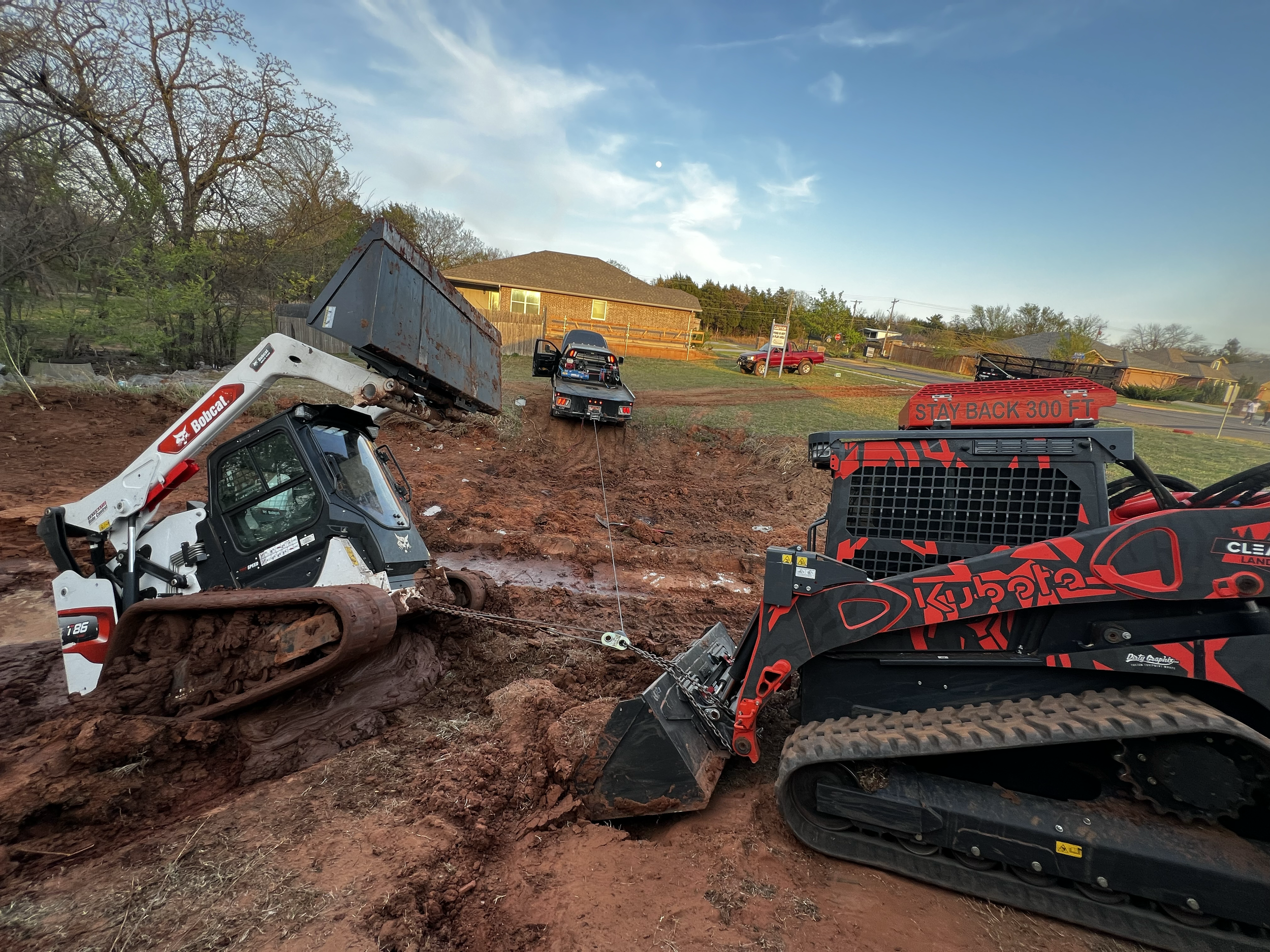 Construction site with machinery, including a Bobcat loader and other vehicles, on dirt in a residential area with houses and trees in the background.