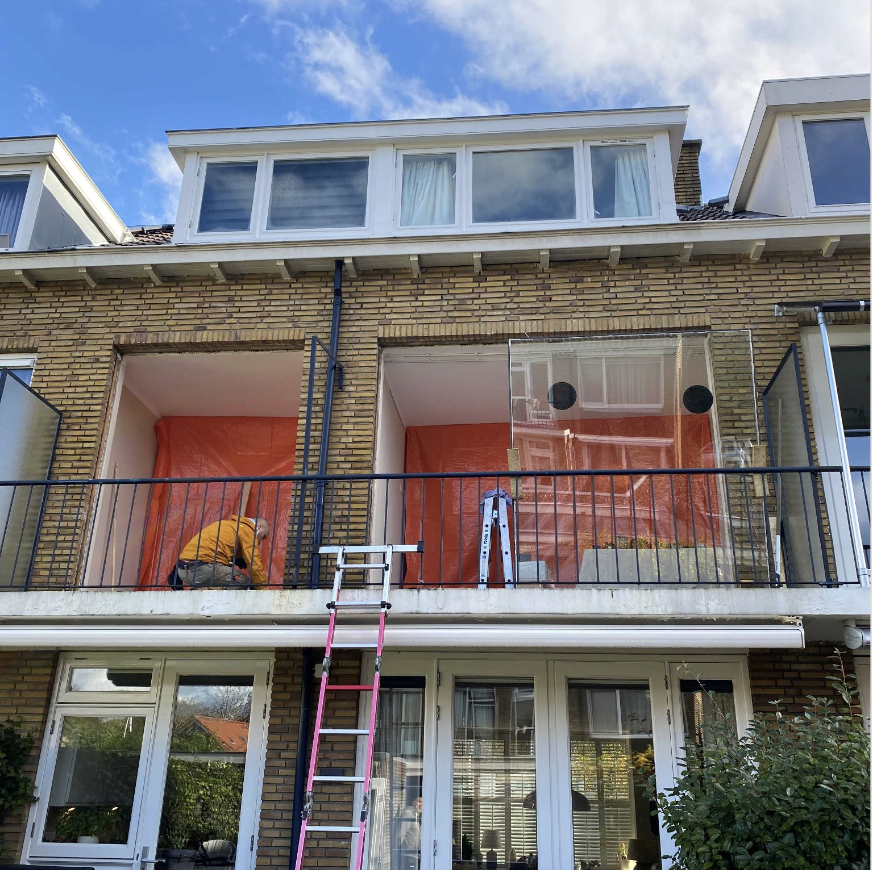 Een man die werkzaamheden op een balkon uitvoert, met een ladder eronder, rond het midden van een brickstenen gebouw, met een blauwe lucht en enkele wolken op de achtergrond.
