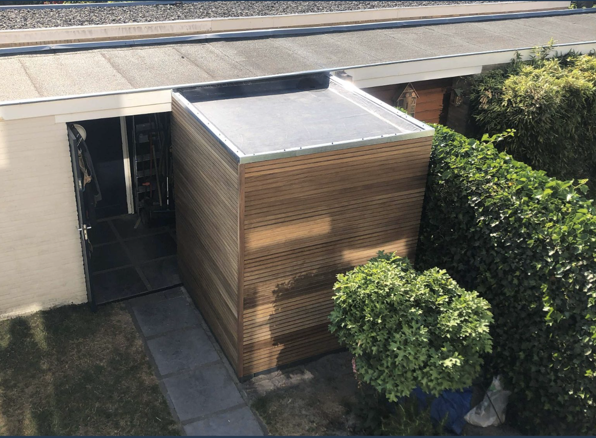 Een houten tuinhuisje naast een woning, met een heg en een boom ernaast.