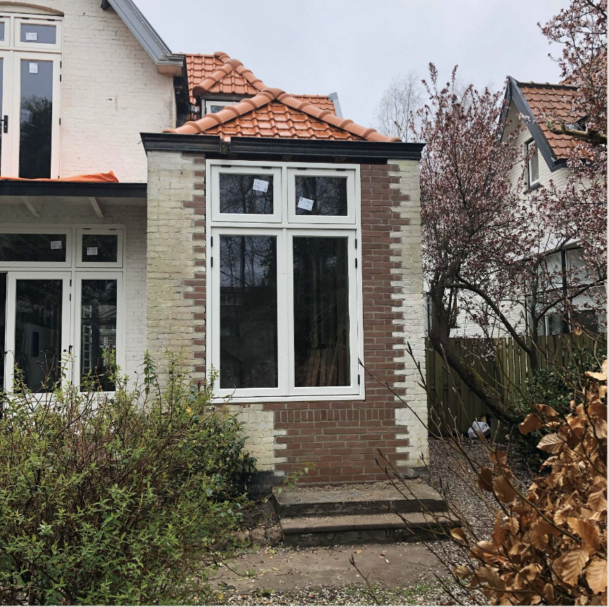 Nieuw aangebouwde steeg met grote ramen en rode dakpannen aan een huis, omgeven door tuingewassen en bomen.