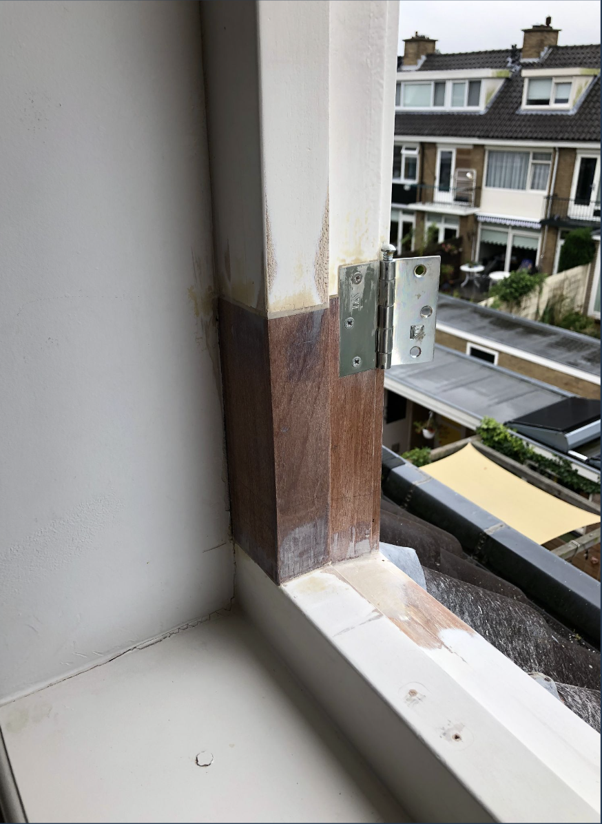 Een close-up van een raamkozijn met een metalen scharnier en een houten steun, zichtbaar vanaf binnenuit met uitzicht op een straat met rowhouses en tuinen.