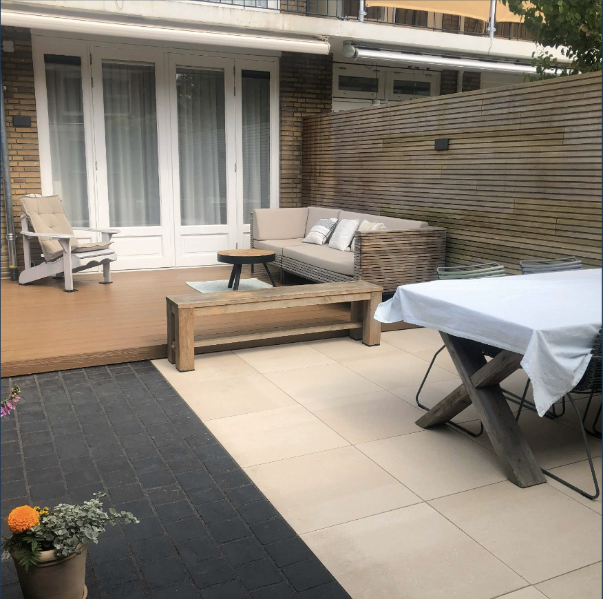 Een buitenruimte met een houten terras met een beige bank, een ronde bijzettafel en een verstelbare ligstoel. Daarvoor een tafel met een witte tafelkleed en enkele stapelbare stoelen. Op de voorgrond een planter met bloemen.