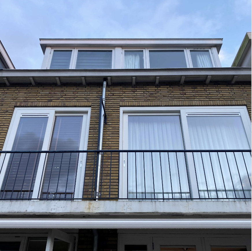 De foto toont de gevel van een appartementengebouw met grote glazen deuren en ramen, een balkon met een zwart metalen hek en een uitbouw met grote ramen aan de bovenverdieping. De gevel is gemaakt van baksteen.