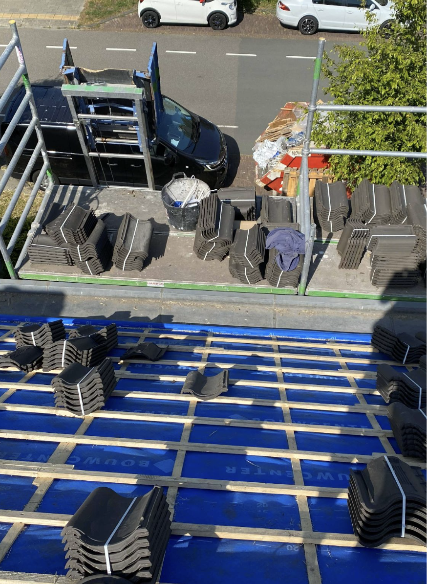 Bovenaanzicht van een bouwplaats met gestapelde dakpannen, een ladder, een schildersdoek, een bak met verfrollers, een grote boom, een geparkeerde auto en bouwmaterialen in de stoep.