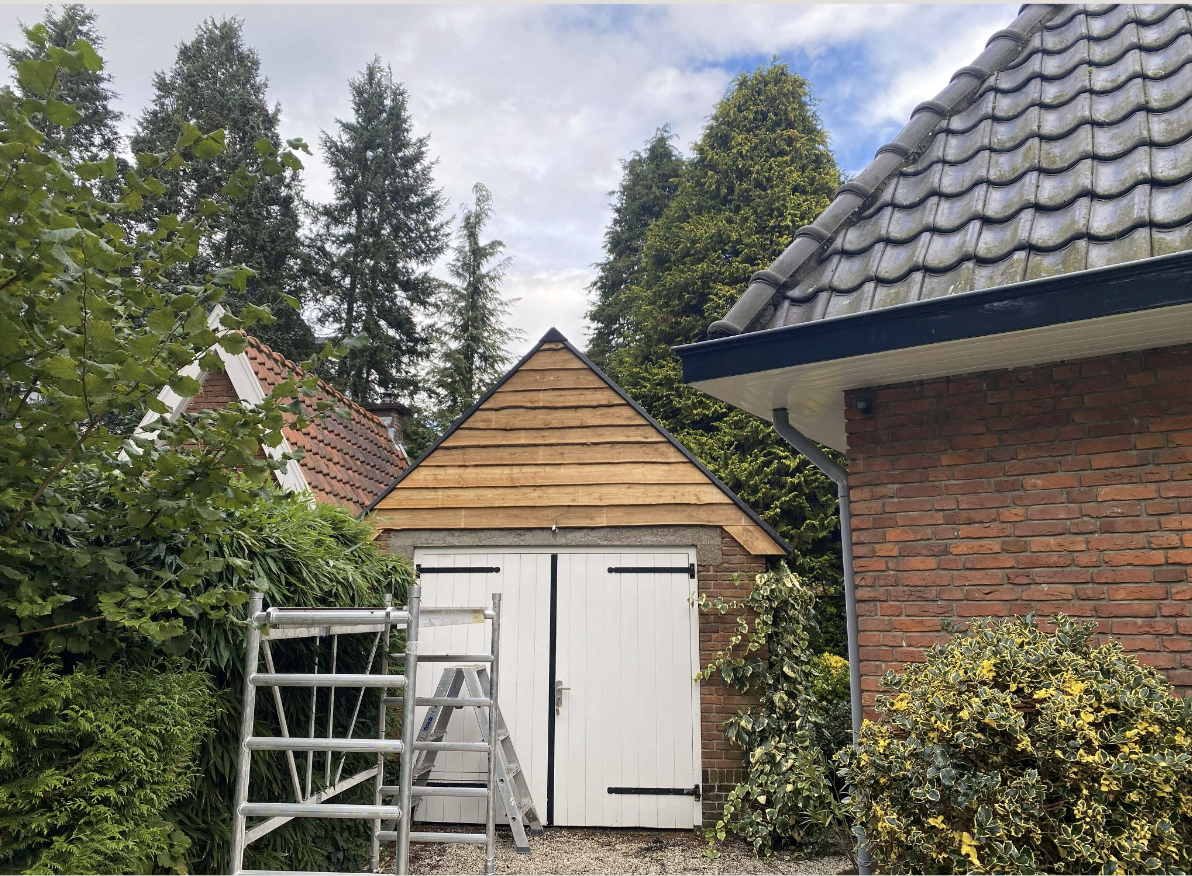 Een tuin met een schuur die in aanbouw is, omgeven door groene bomen en planten. Van links naar rechts zijn er grote struiken en een ladder voor de werkzaamheden. Rechts is een deel van een huis te zien met een dak met golvende dakpannen en een gotische rand, gemaakt van rood baksteen.
