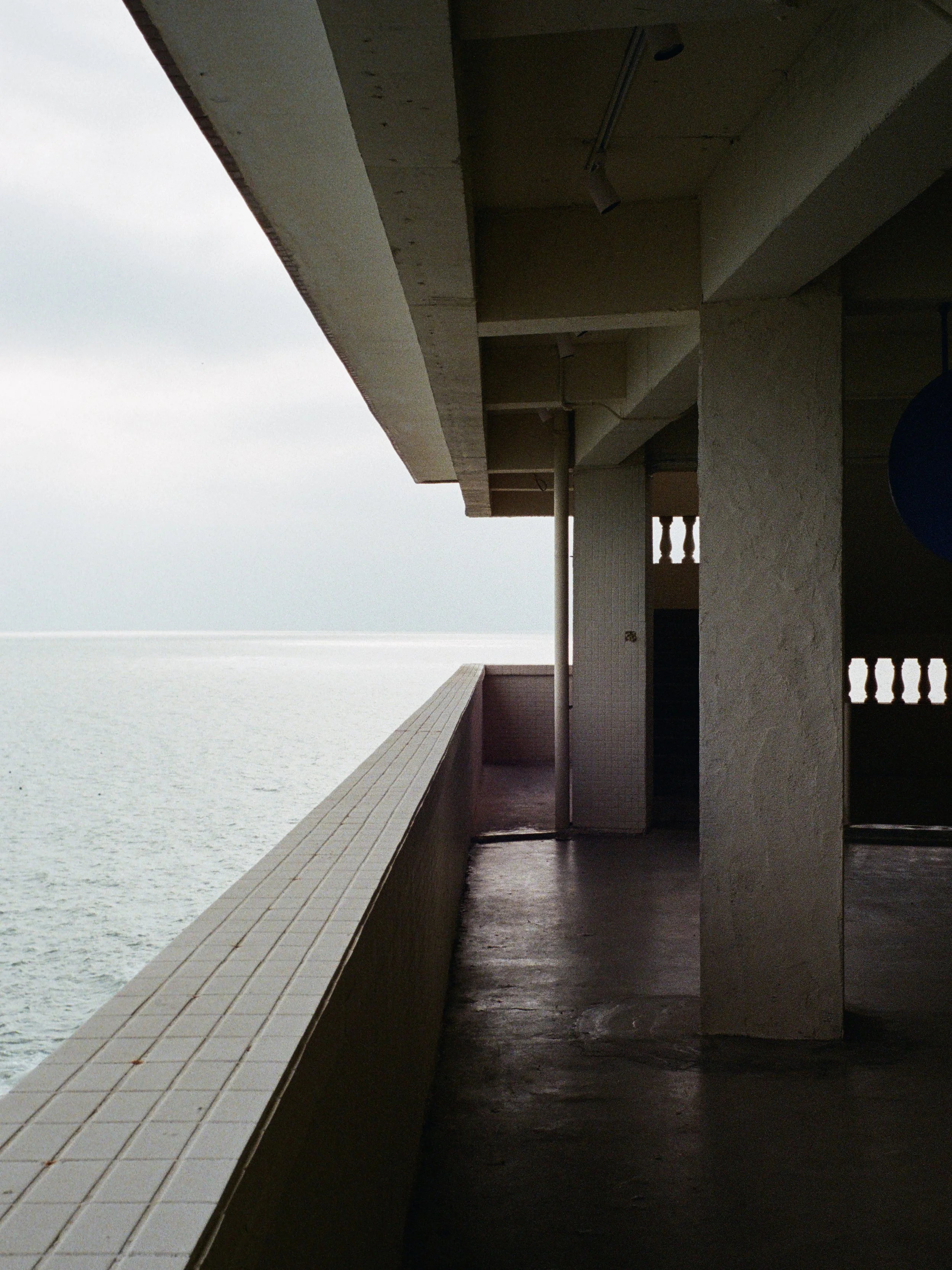 Empty balcony with white tiled floor, beige walls, and a railing overlooking the ocean. Overcast sky.