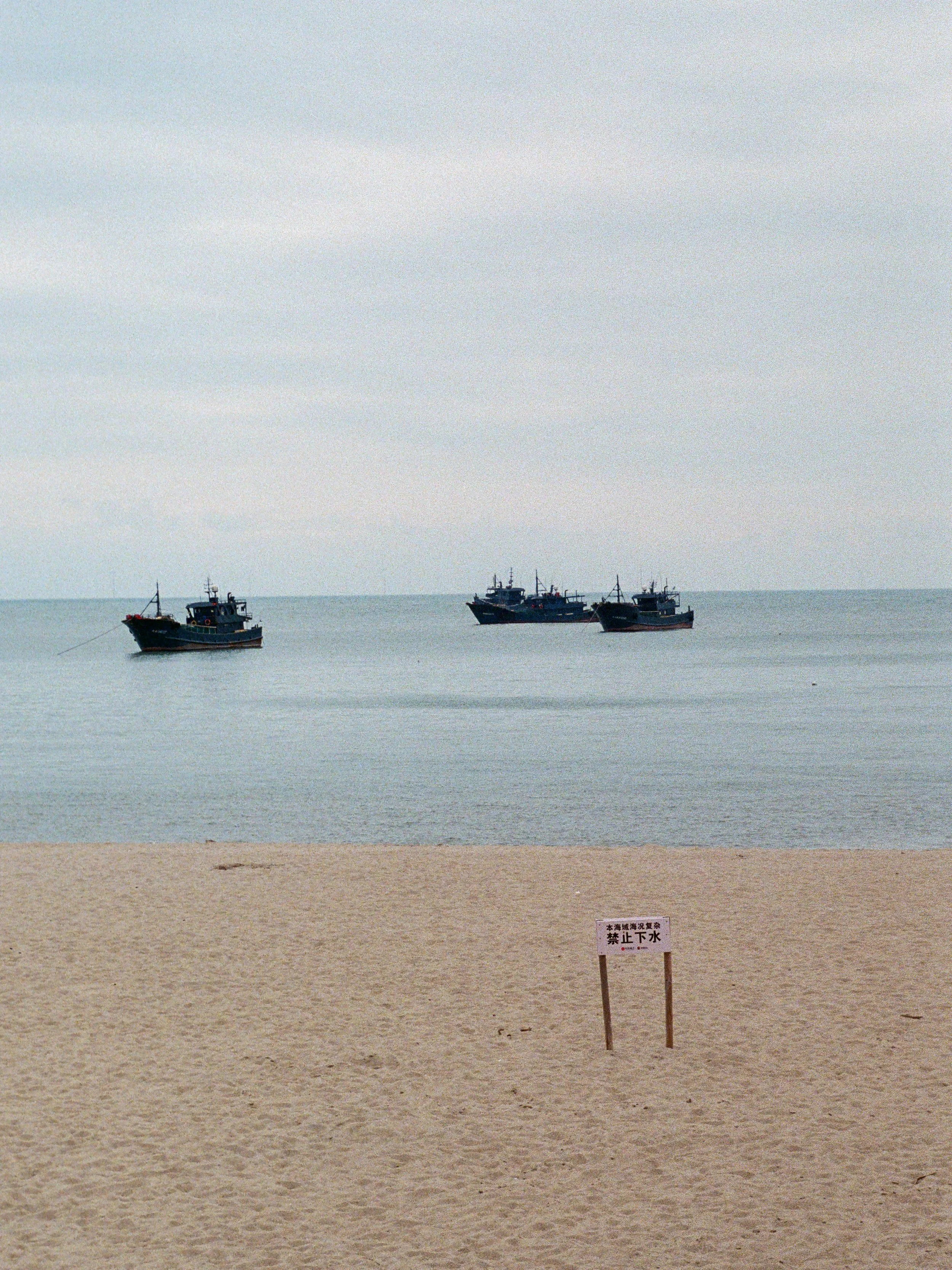 A beach with a sandy shore, calm water, and four small boats floating in the water. There is a sign on the beach with Japanese writing.