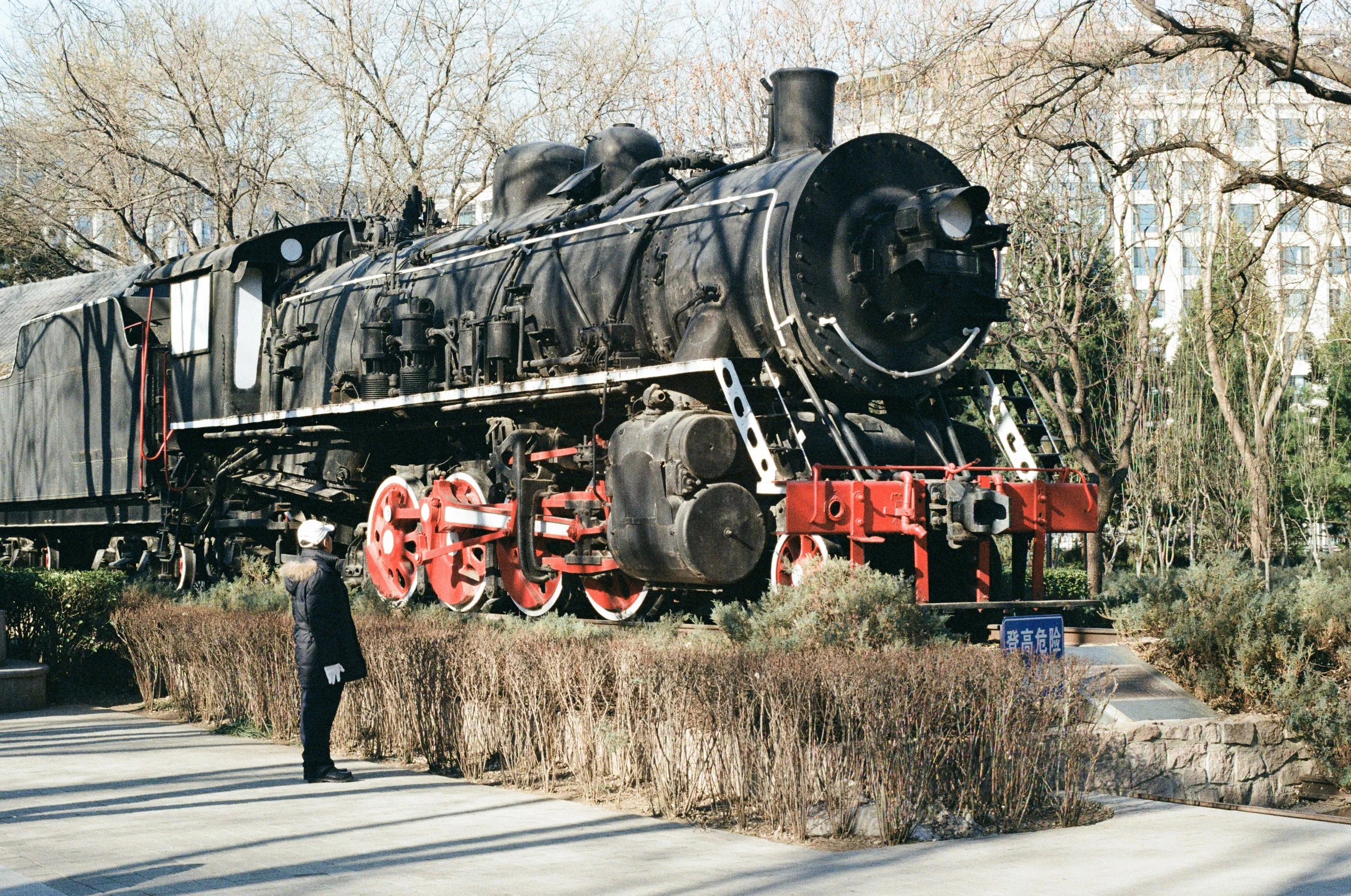 A vintage black steam locomotive on display outdoors, with a person standing nearby and trees in the background.