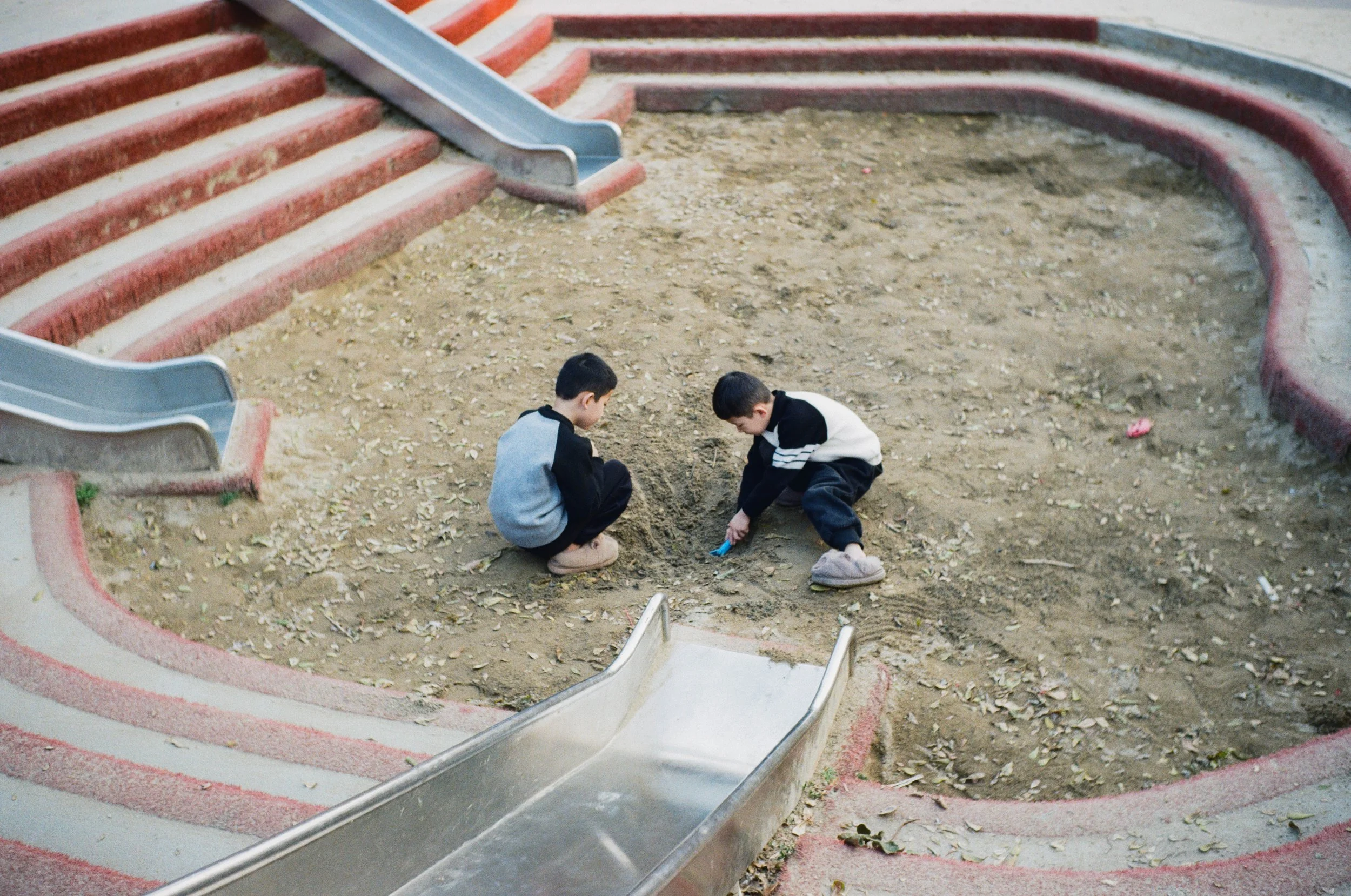 Two boys playing in a dugout sandpit area at a playground, sitting on the ground with tools.