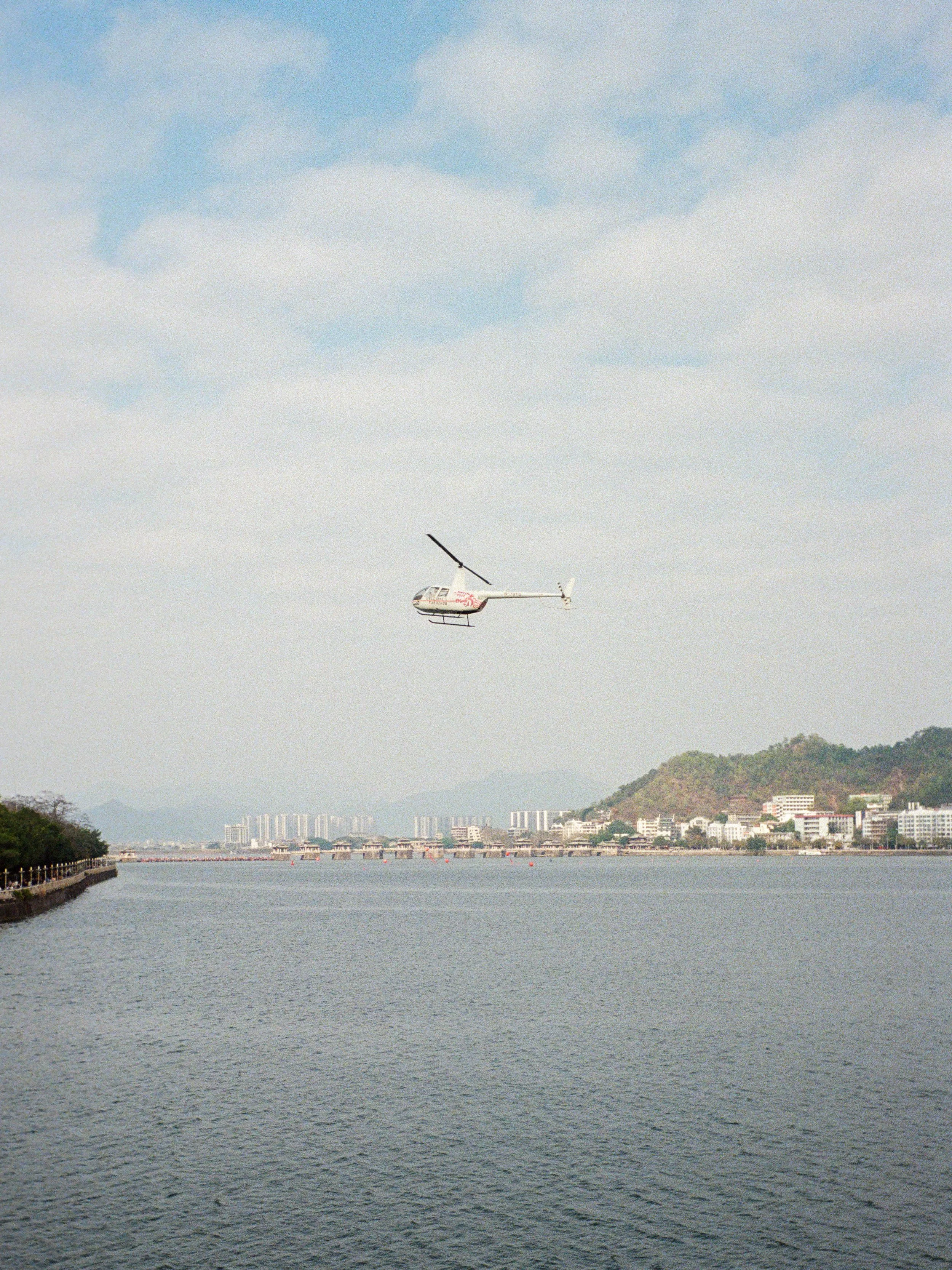A helicopter flying over a body of water near a city with mountains in the background and a partly cloudy sky.