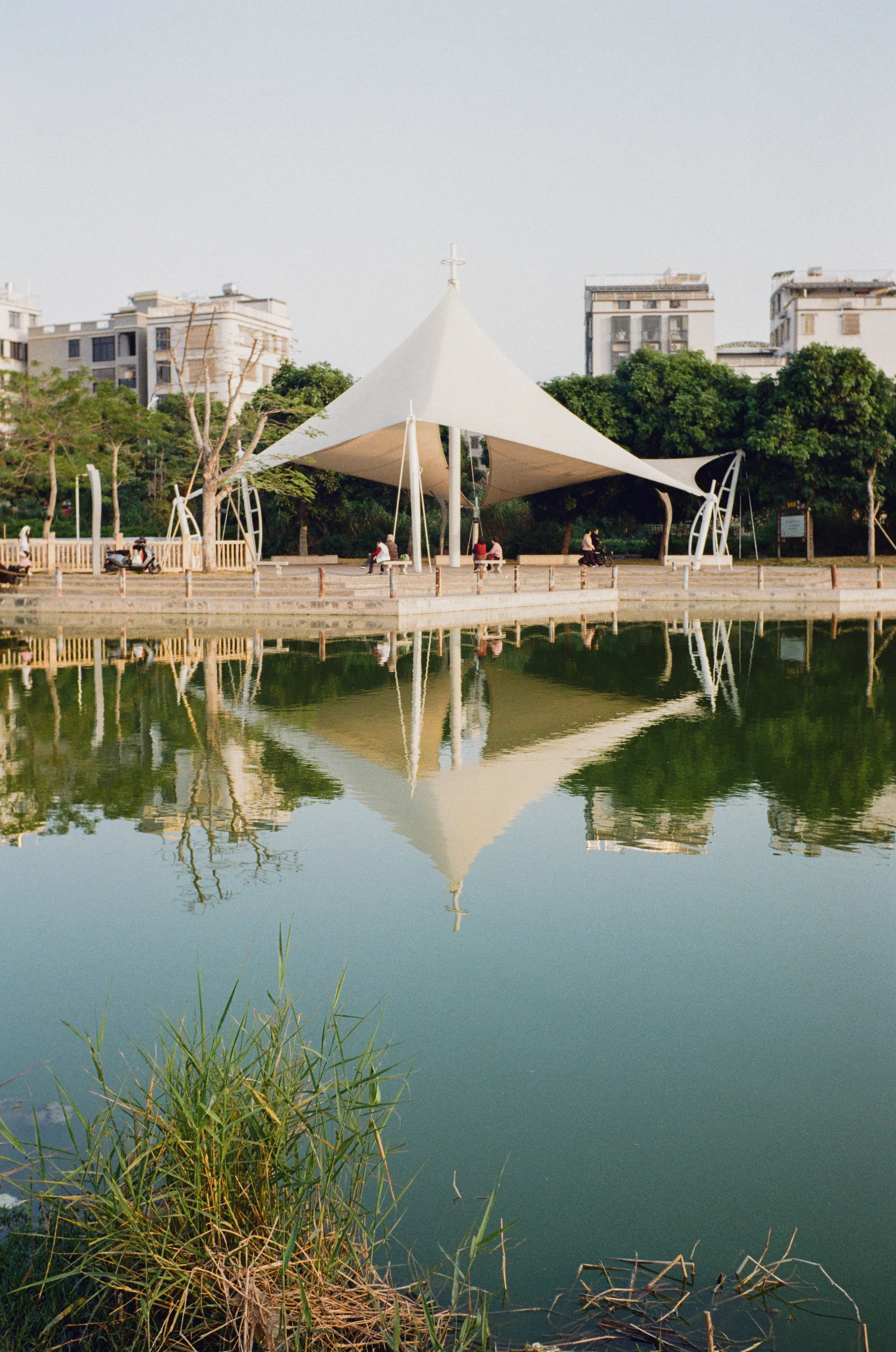 A white, tensioned fabric canopy structure with a pointed peak and a cross on top, located over a park with trees and benches. There is a small body of water in the foreground reflecting the structure and surroundings, with grass and plants at the wa