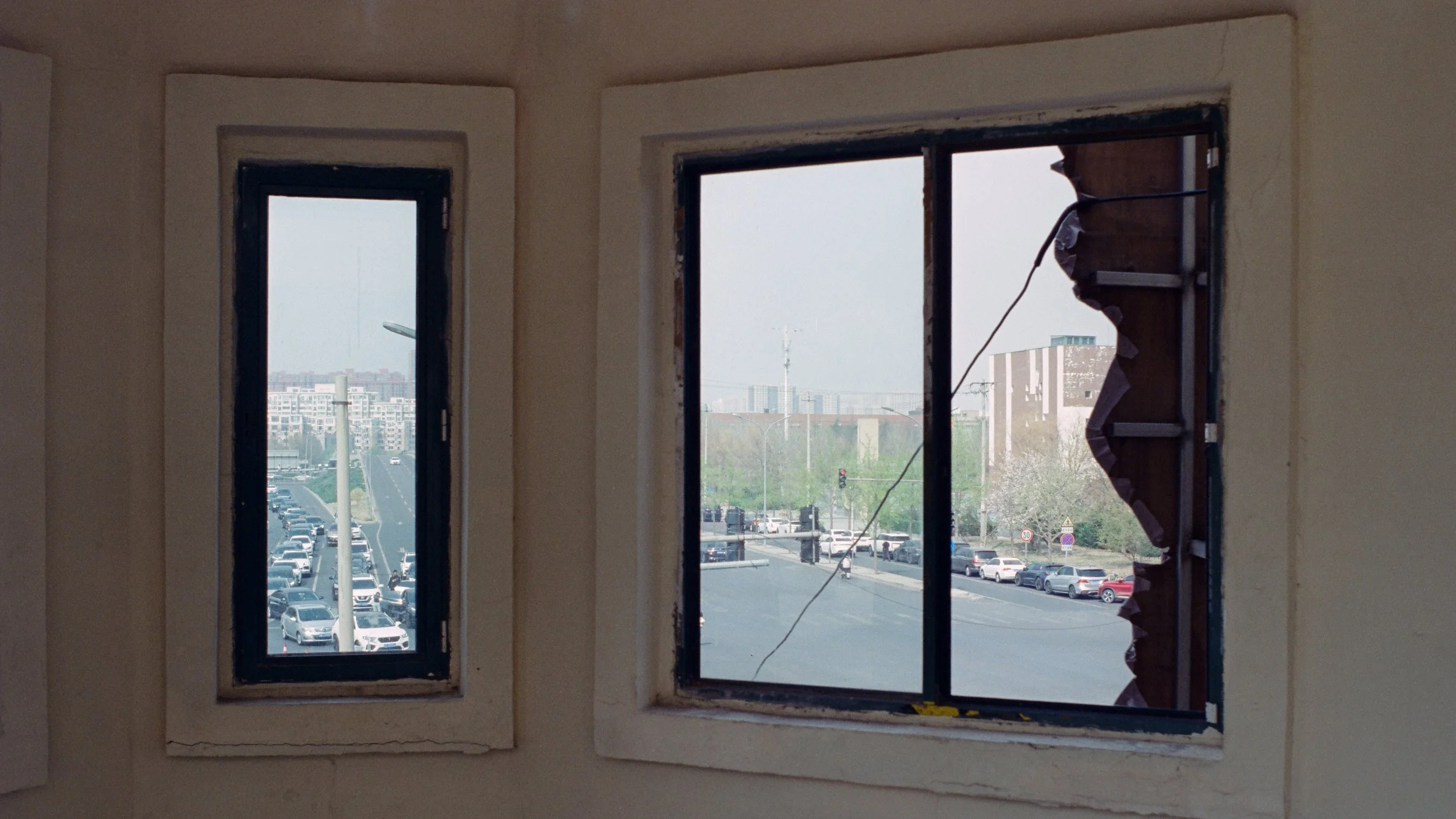 Three windows in a wall, with one broken and a view of a busy parking lot and cityscape outside.