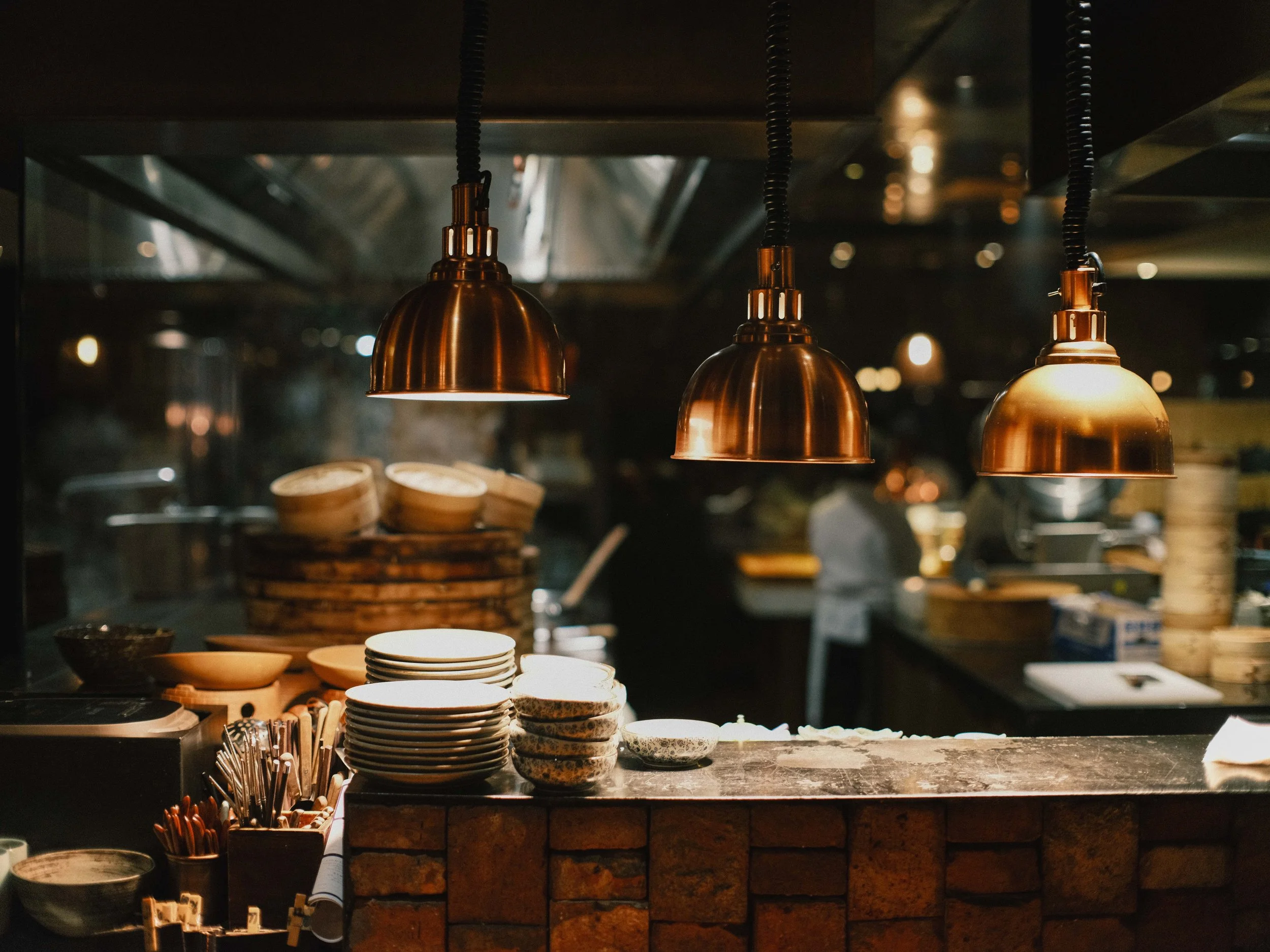 Copper pendant lights hanging above a counter with stacked plates, bowls, and utensils in a restaurant or cafe setting.