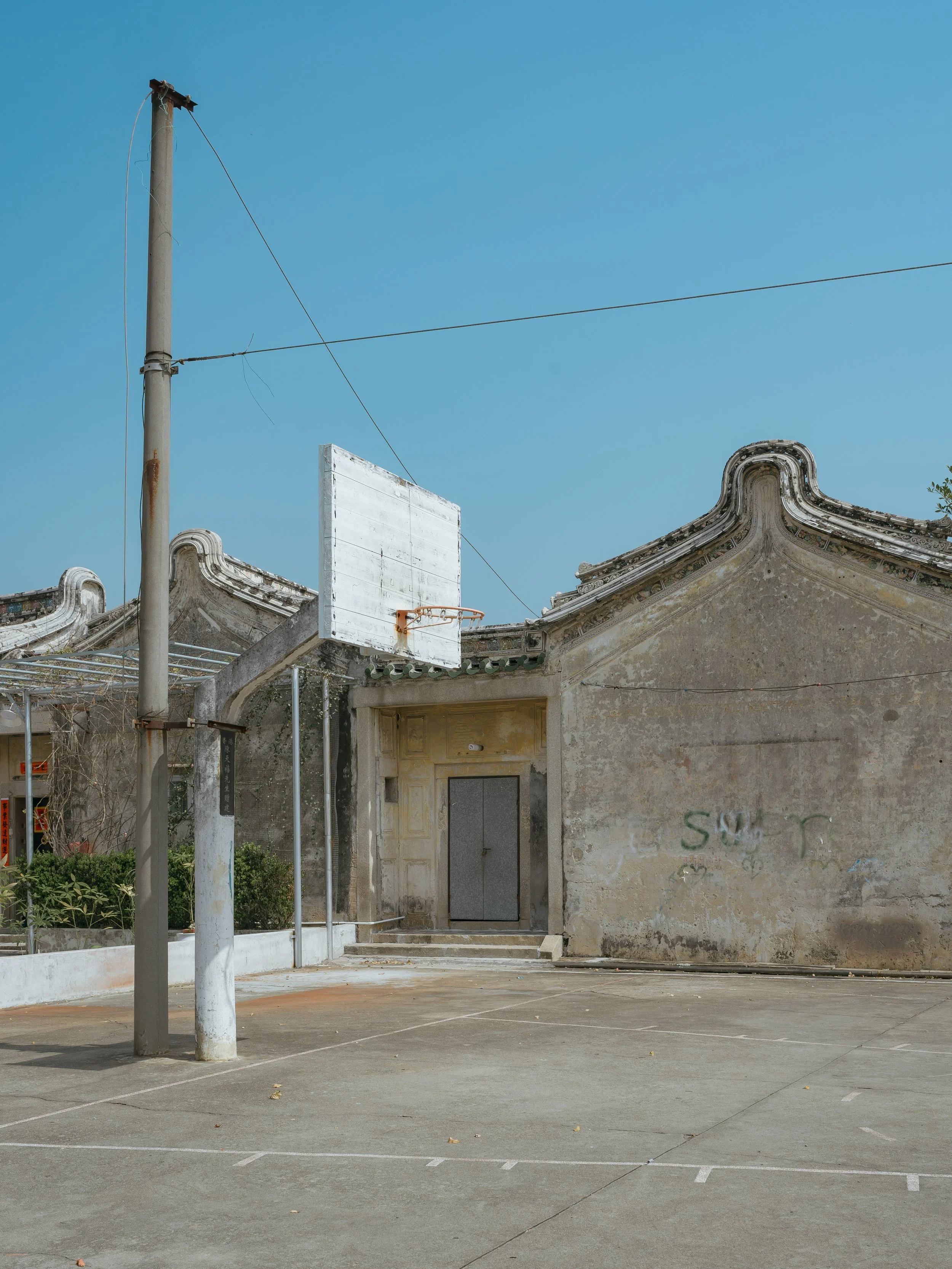 An empty outdoor basketball court with a building in the background, a utility pole with wires, and a basketball hoop without a net.