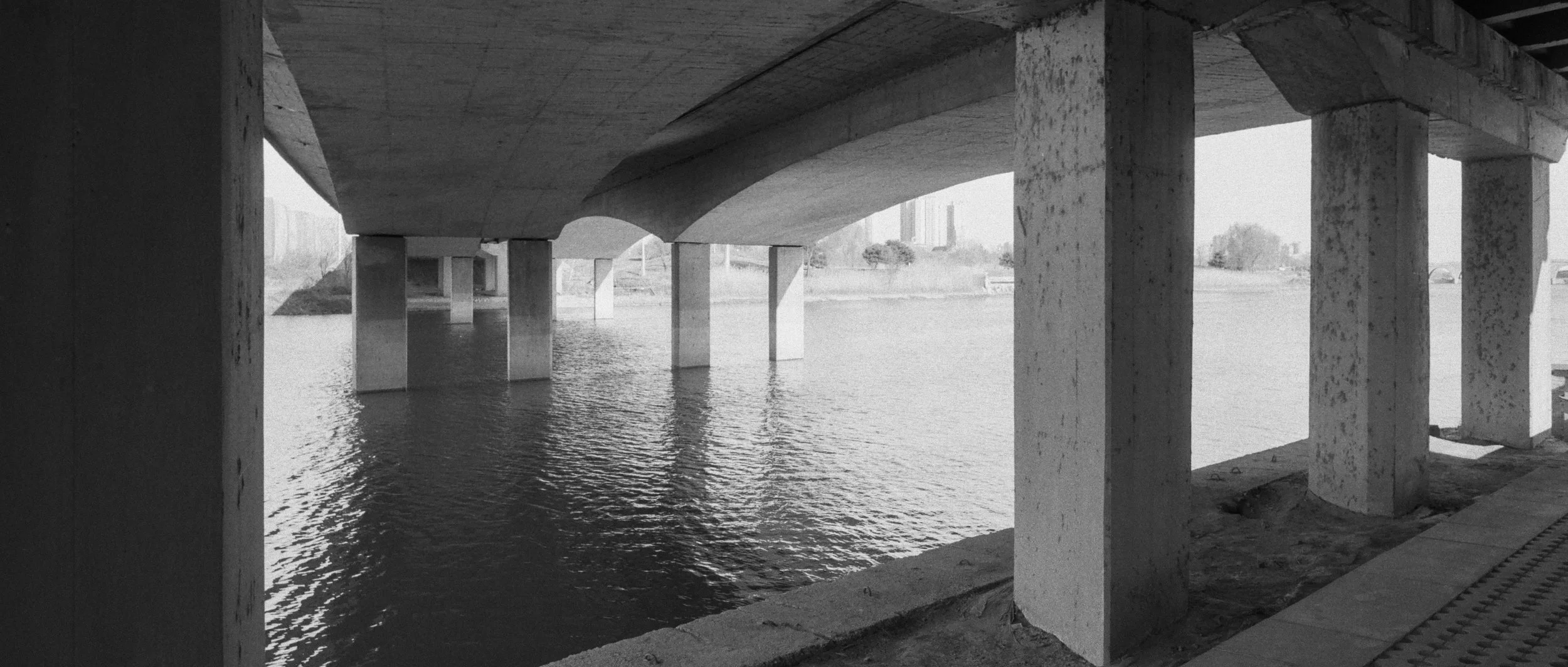 View of a bridge's underside and support pillars over a body of water, with a cityscape in the background.