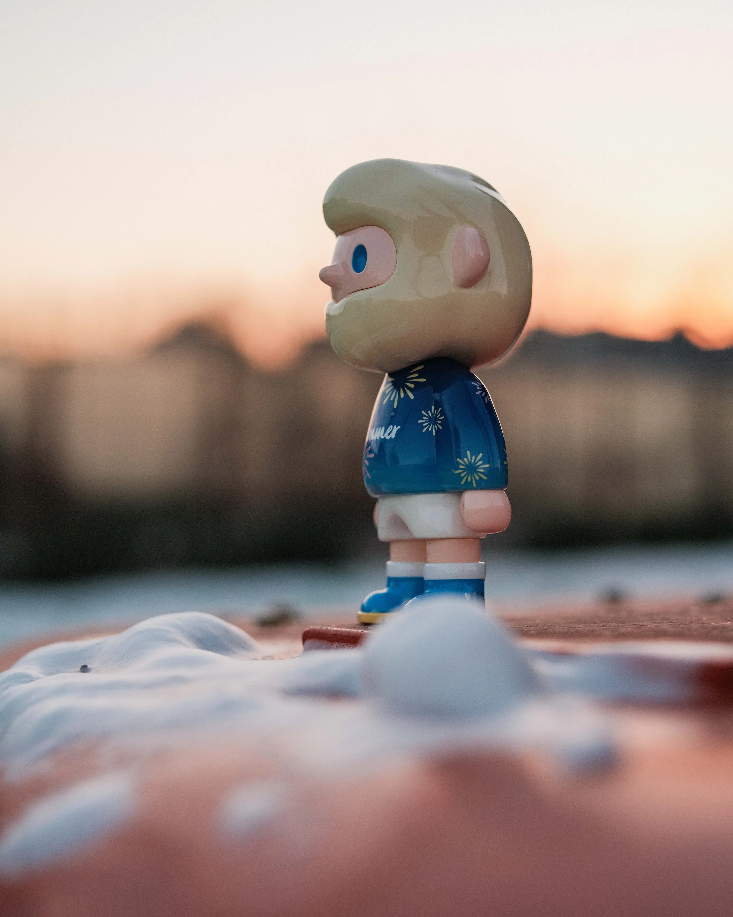 A small figurine with a girl character, blue eyes, and white hair, wearing a blue dress with yellow flowers, standing on a surface with snow or snow-like material, with a blurred sunset sky in the background.