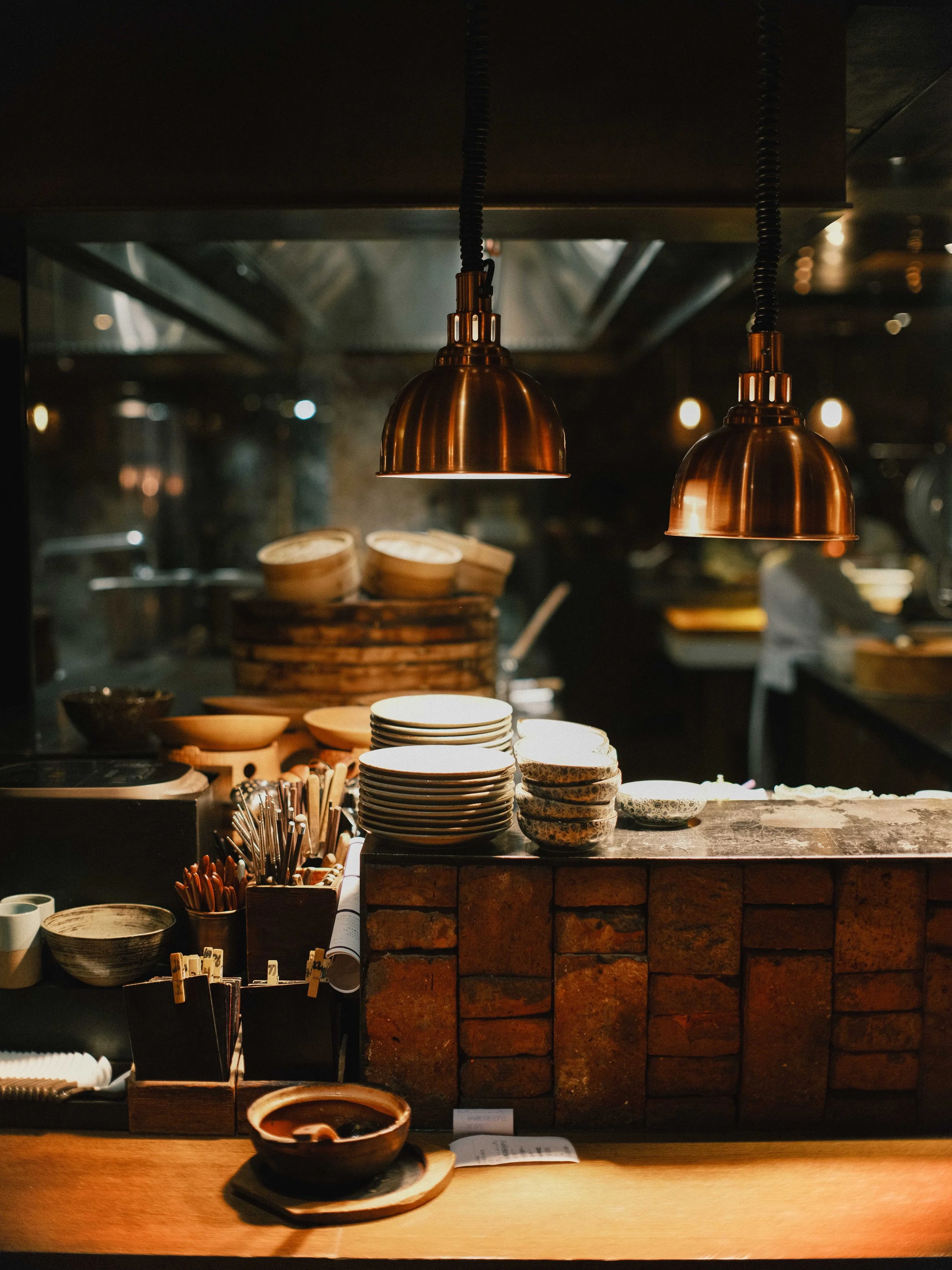 A kitchen counter with stacked bowls, chopsticks, and a ceramic bowl in front. Behind, shelves with more bowls and kitchen utensils, under warm hanging lights.