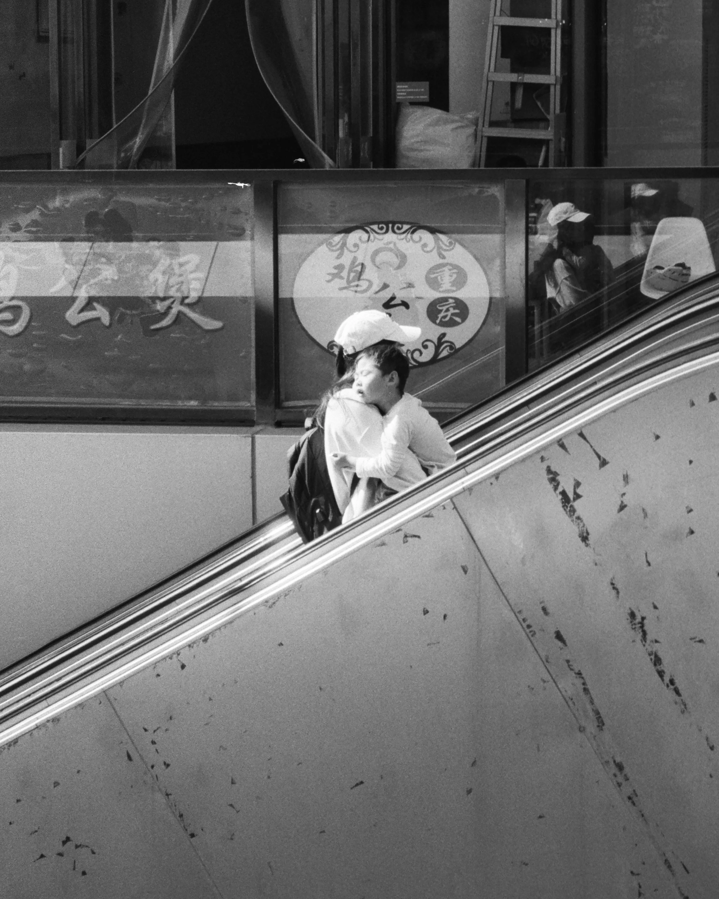 Black and white photo of a hand holding a baseball cap, standing in front of a storefront with Chinese characters on sign, reflected in glass window. A person in a jacket and cap is also reflected in the window, with an escalator at the bottom of the