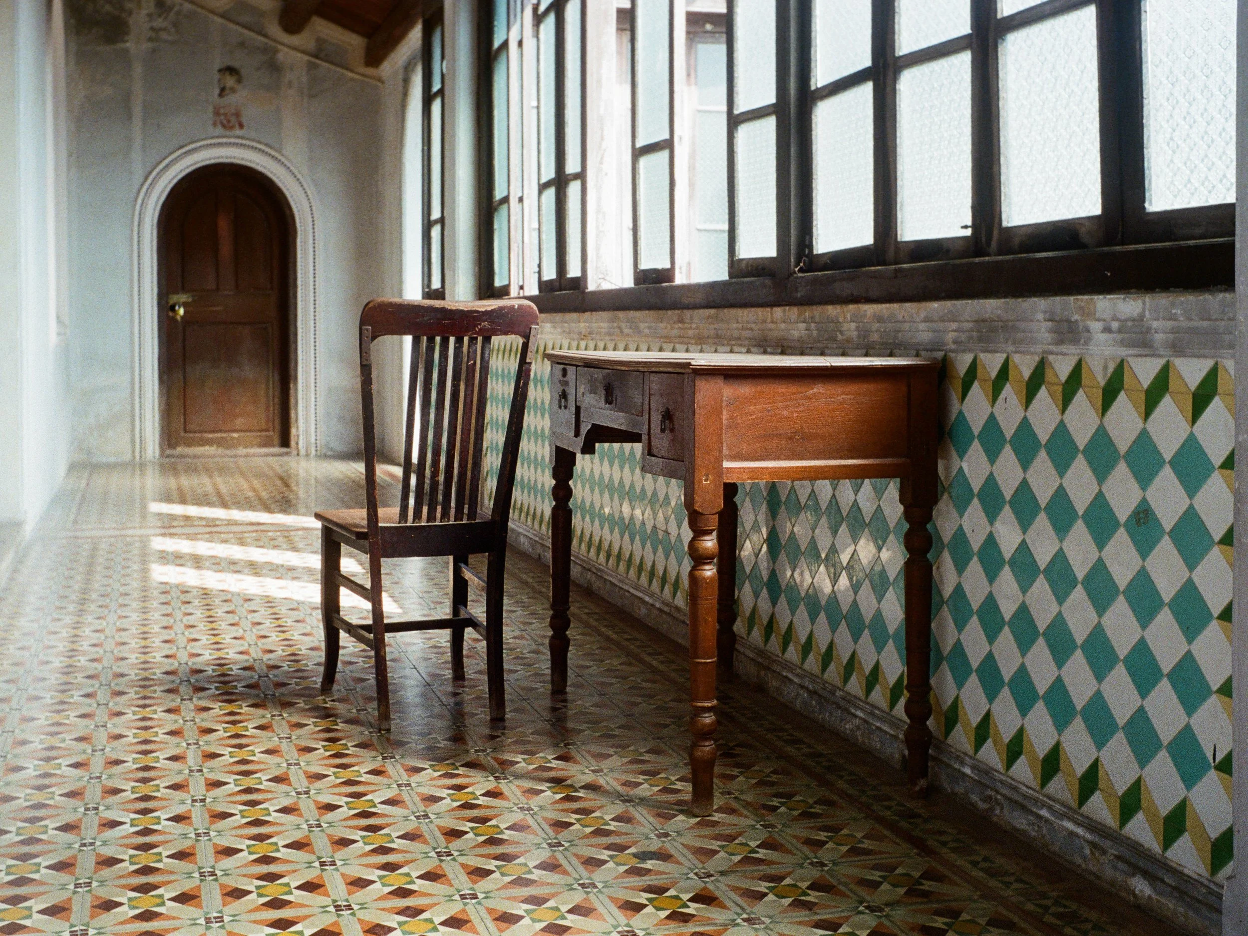 Interior of a historic building with patterned tile flooring, a wooden chair, a small wooden desk, a set of large windows, and a door in the background.