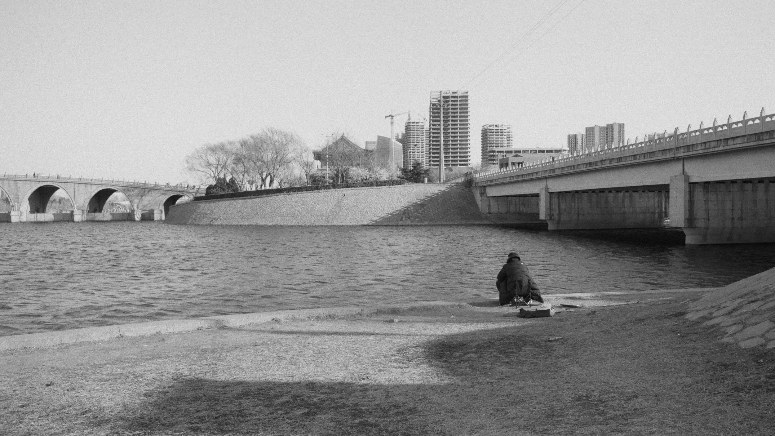 A person sitting on the edge of a body of water, possibly fishing, near a cityscape with high-rise buildings and a bridge in the background.