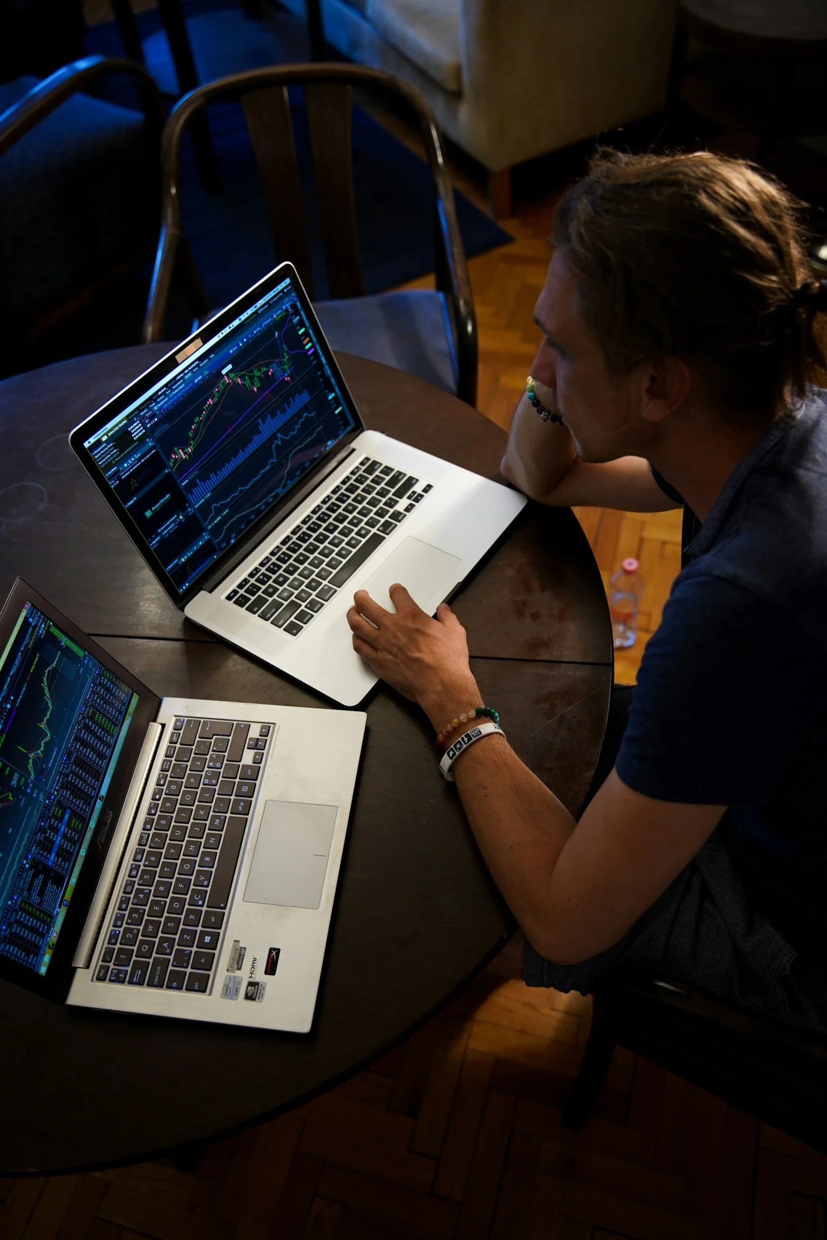 A person sitting at a table in a dimly lit room, working on a laptop with a financial chart on the screen. There are two laptops on the table, one open with a colorful stock or forex trading chart, and another partially visible. The person appears focused, resting their head on one hand, with a black bracelet and beaded bracelet on their wrist.