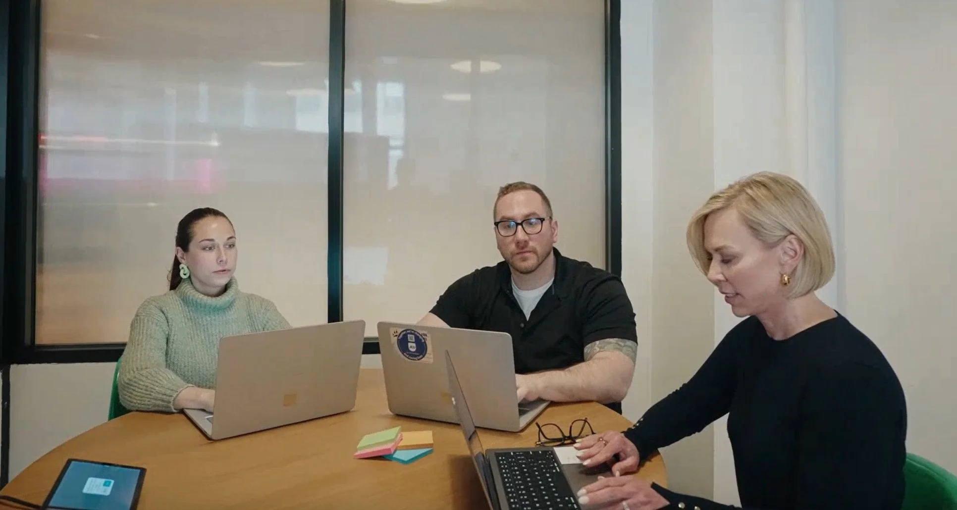 Three people sitting at a table with laptops in a meeting room, two women and one man, focused on their work.