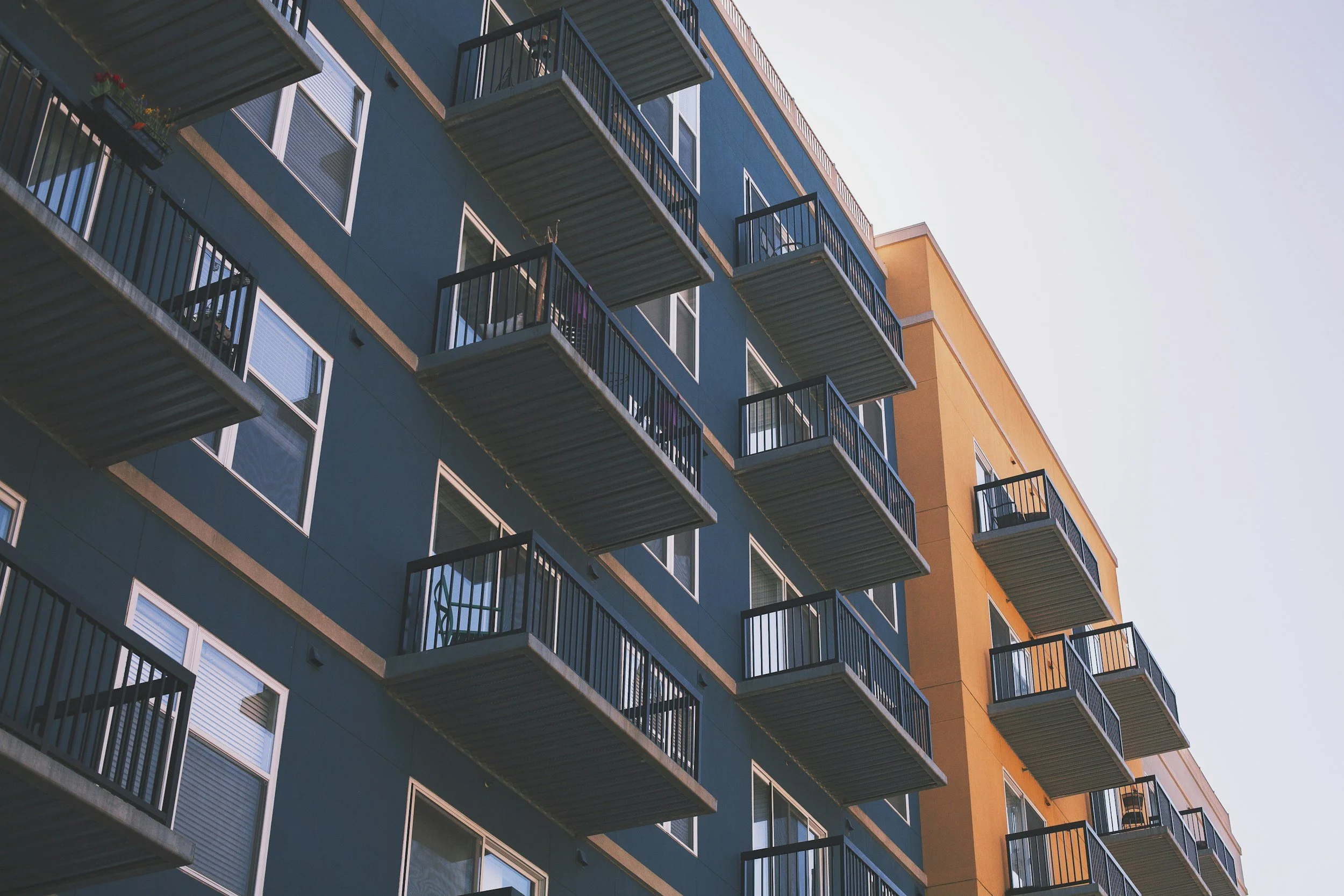 A tall apartment building with a blue exterior, orange section, and multiple balconies with black railings against a clear sky.