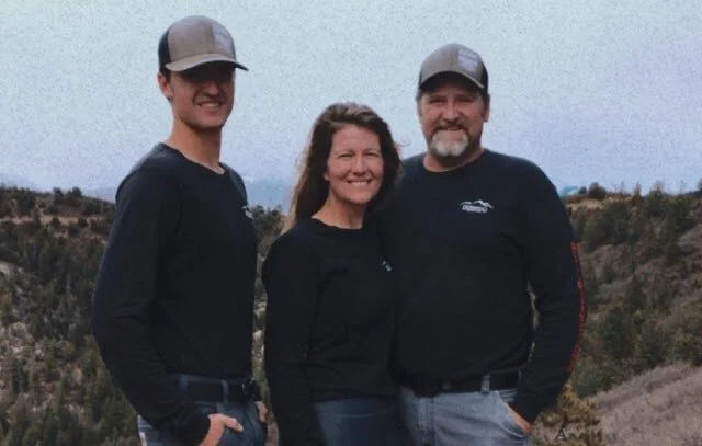 Three people standing outdoors in a hilly area, smiling at the camera. Two men on either side and a woman in the middle.