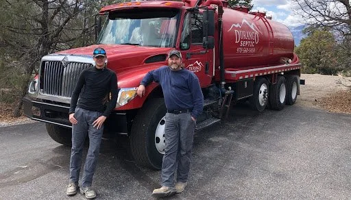 Two men standing in front of a large red Durnago septic truck on a paved road with trees and cloudy sky in the background.