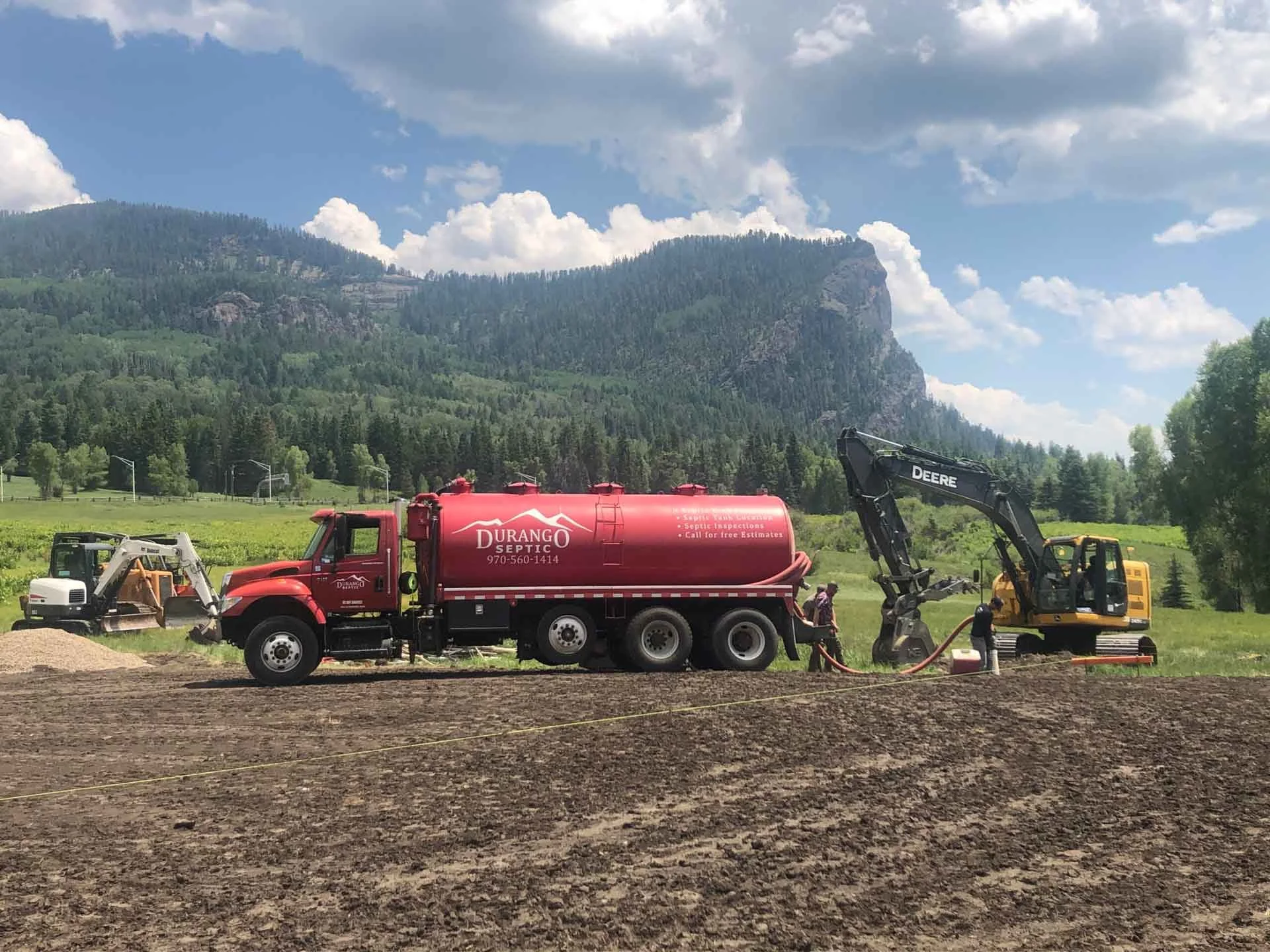 A red Durango septic truck parked on site delivering septic services with a yellow Deere excavator equipment behind it.