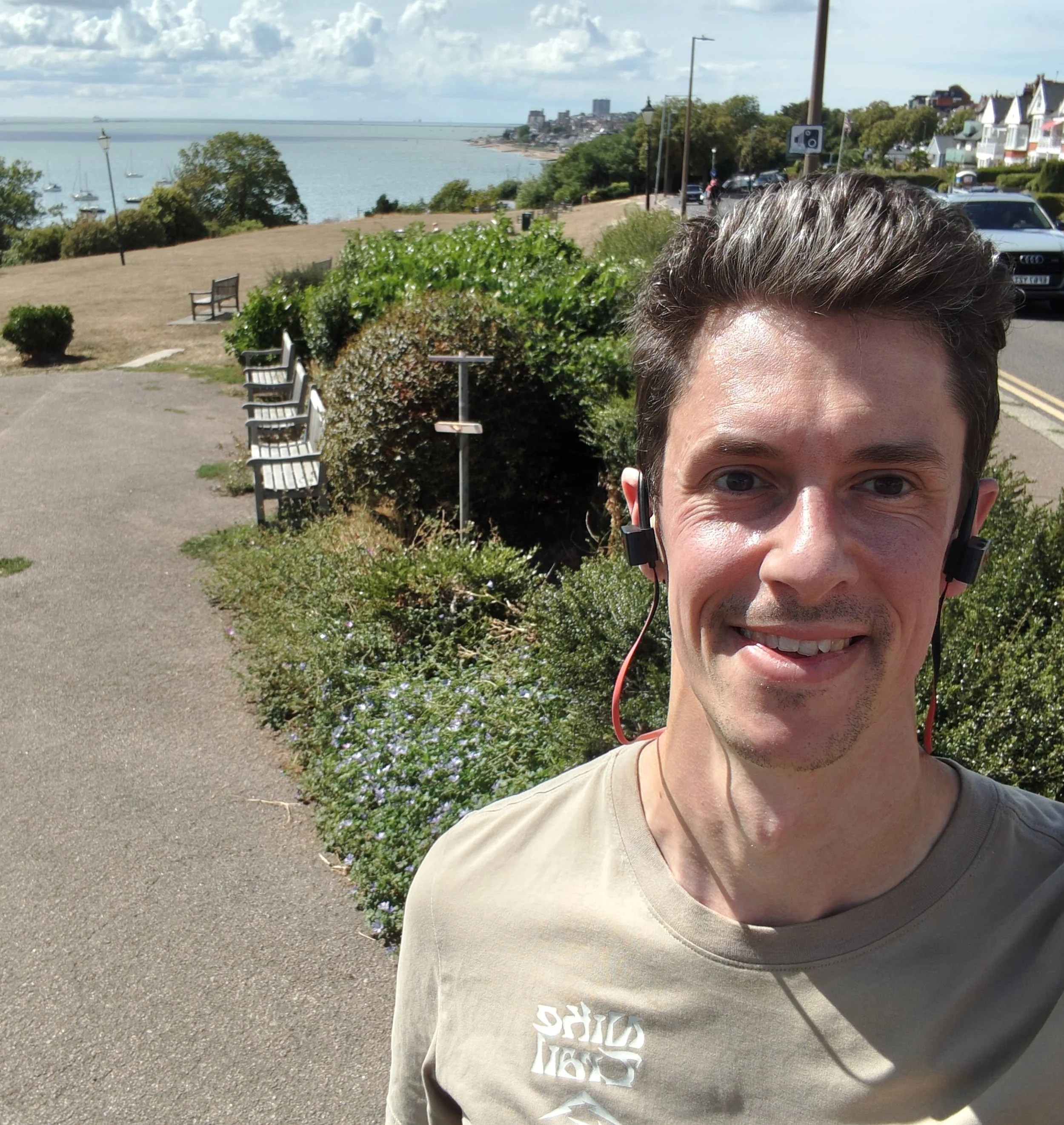 Close-up of a smiling man wearing a beige T-shirt and headphones, with an estuary, sailboats and trees in the background.
