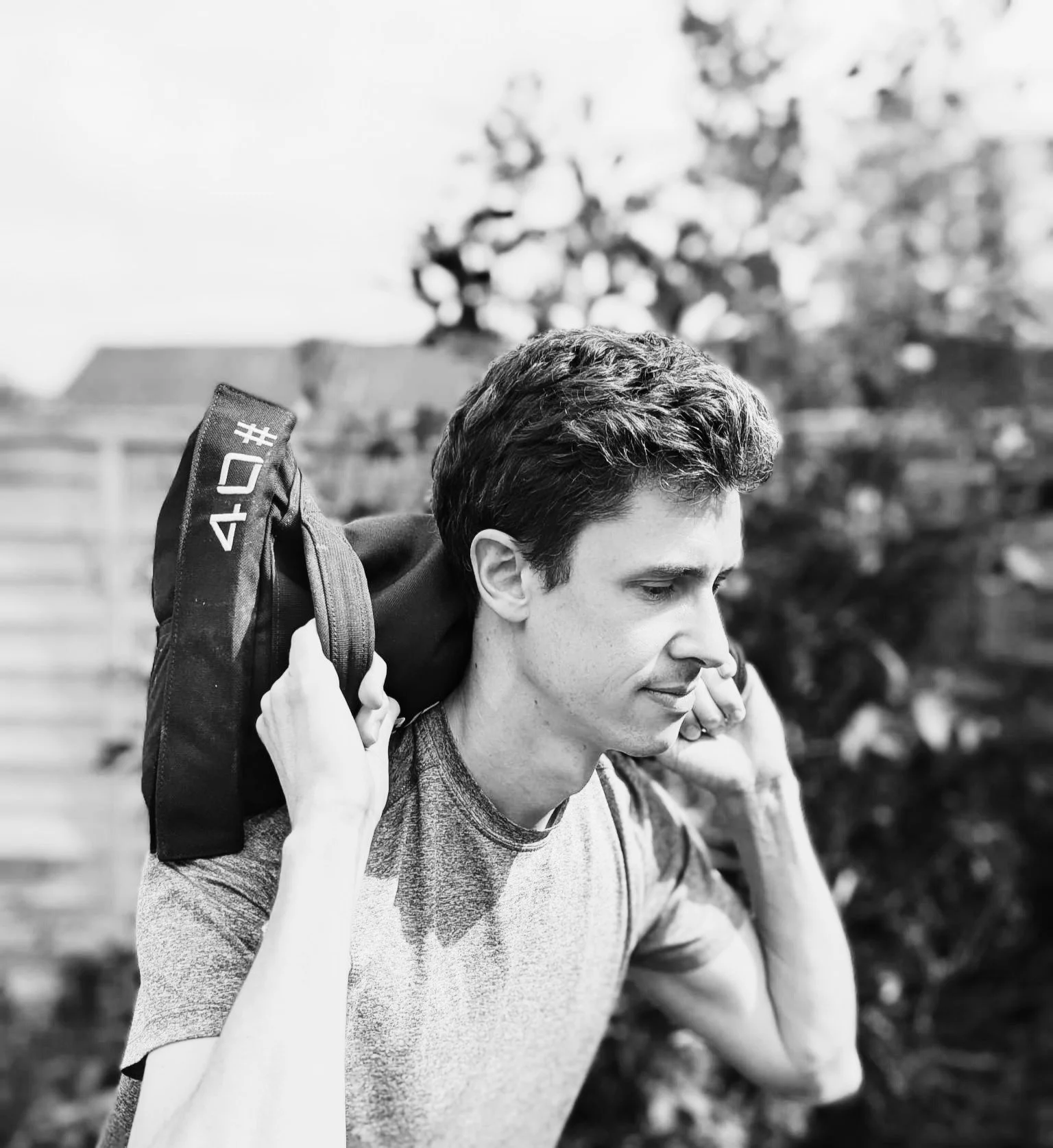 A young man with dark hair squatting whilst holding a black sandbag across his shoulders, outdoors with trees and a fence in the background.