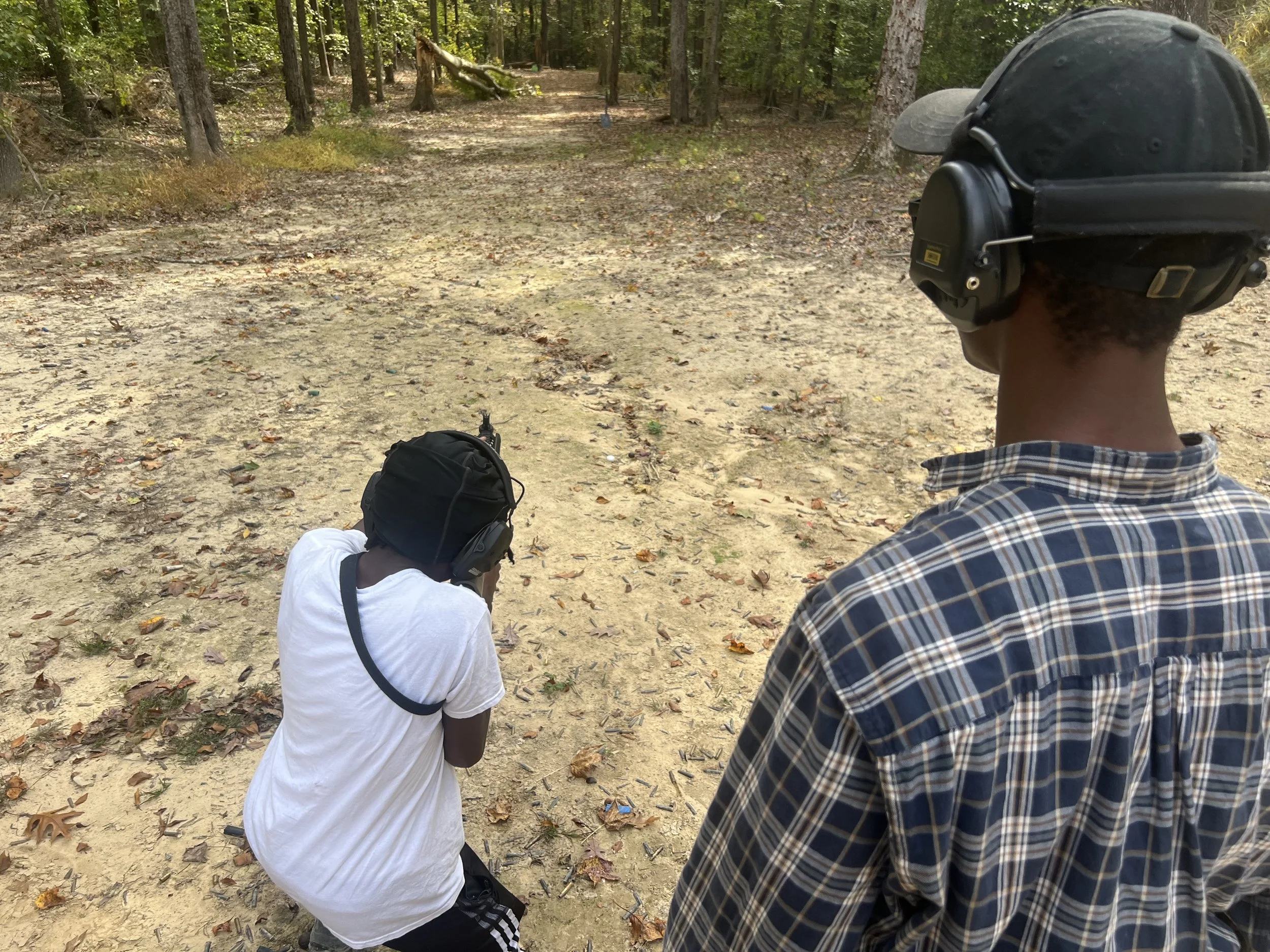 Two children and an adult in a wooded area, all wearing hearing protection, aiming guns at a shooting range.