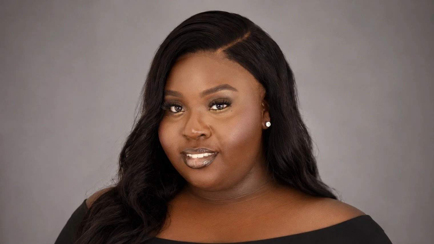 A young African American woman with long wavy black hair, wearing a black off-the-shoulder top and stud earrings, posing against a plain gray background.