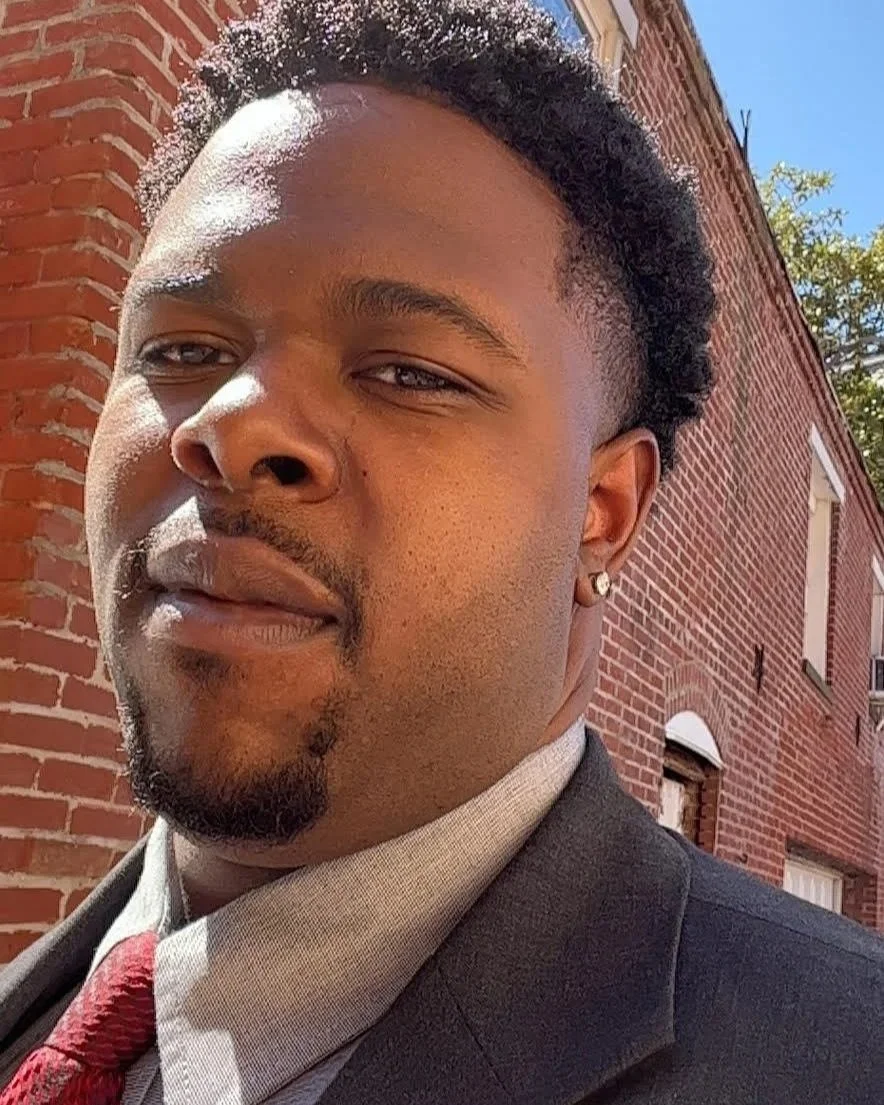 Close-up of a young man outdoors in front of a brick building, wearing a suit and red tie, with sunlight on his face.
