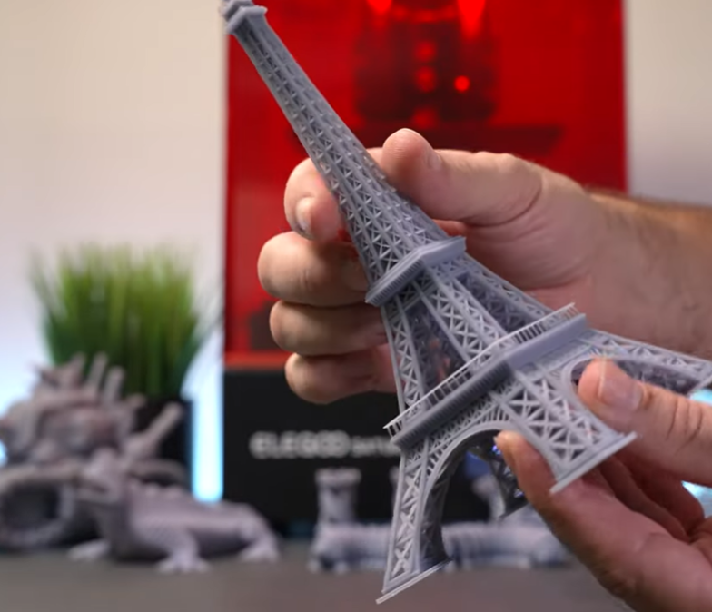 Person holding a gray 3D-printed model of the Eiffel Tower with plants and 3D printer in the background.