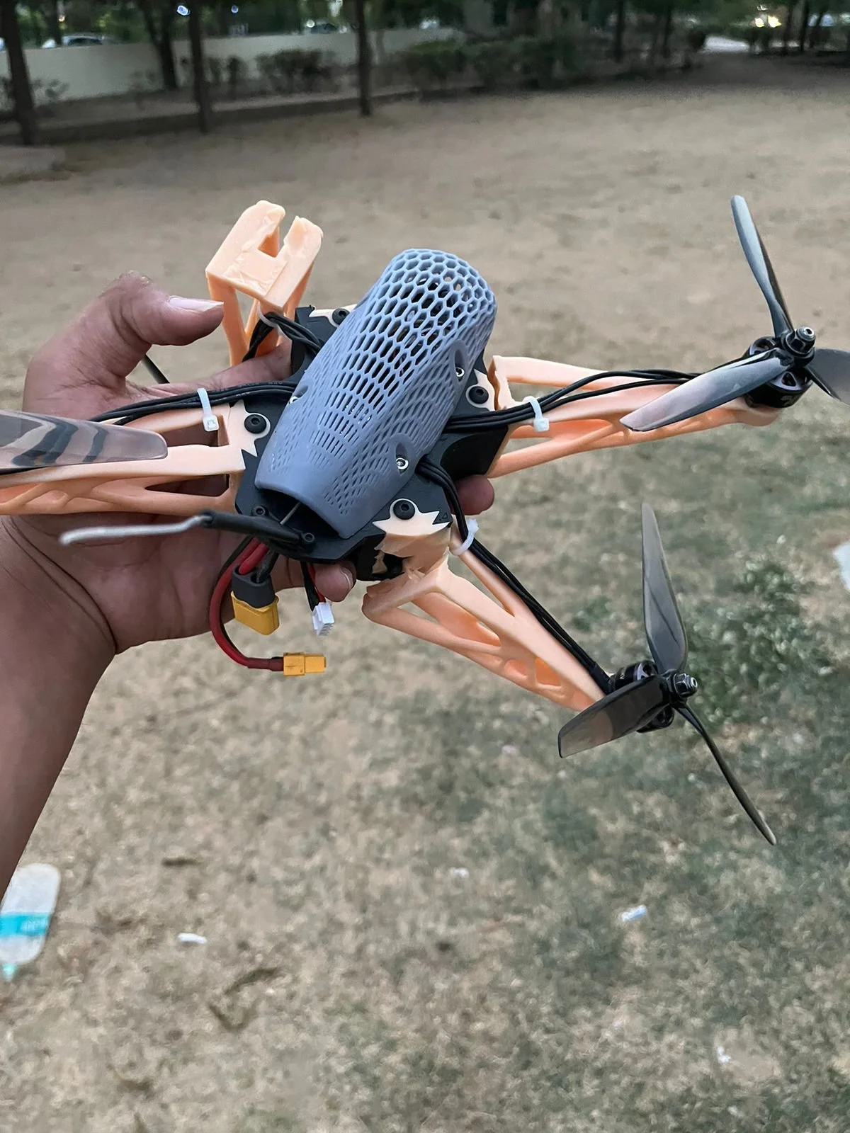 A person holding a small drone with a protective blue cover over the motor and propellers, taken outdoors on a dirt surface.