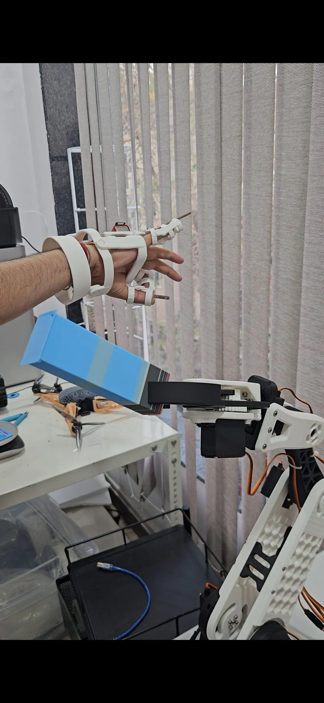 Close-up of a robotic arm holding a human hand with a glove, attached to a mechanical setup on a workstation. The background shows window blinds with a view outside.