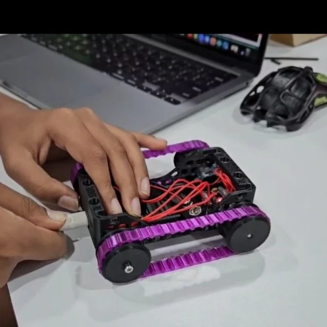 A person assembling a small robot with purple treaded wheels and red wires, with a laptop and a black object on the table nearby.