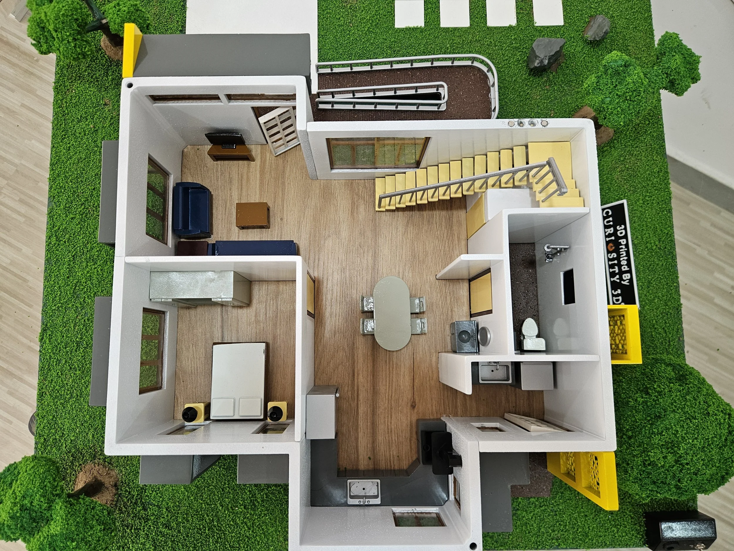 Top-down view of a dollhouse with a living room, kitchen, dining area, bathroom, and bedroom, surrounded by green grass and trees.