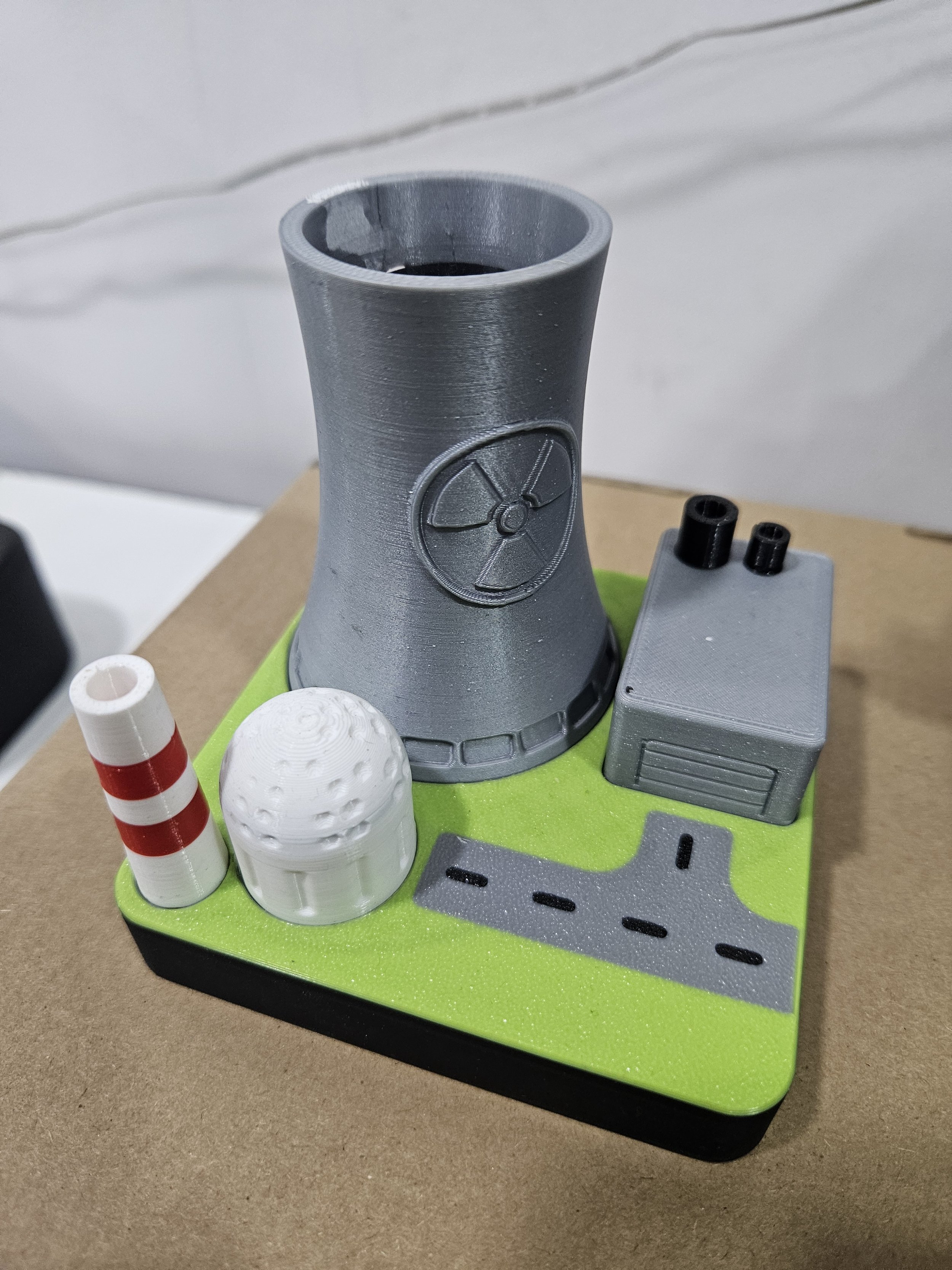 A miniature model of a city scene with a gray tower, a white and red striped traffic cone, a white spherical structure, a gray building, and a road on a green base.