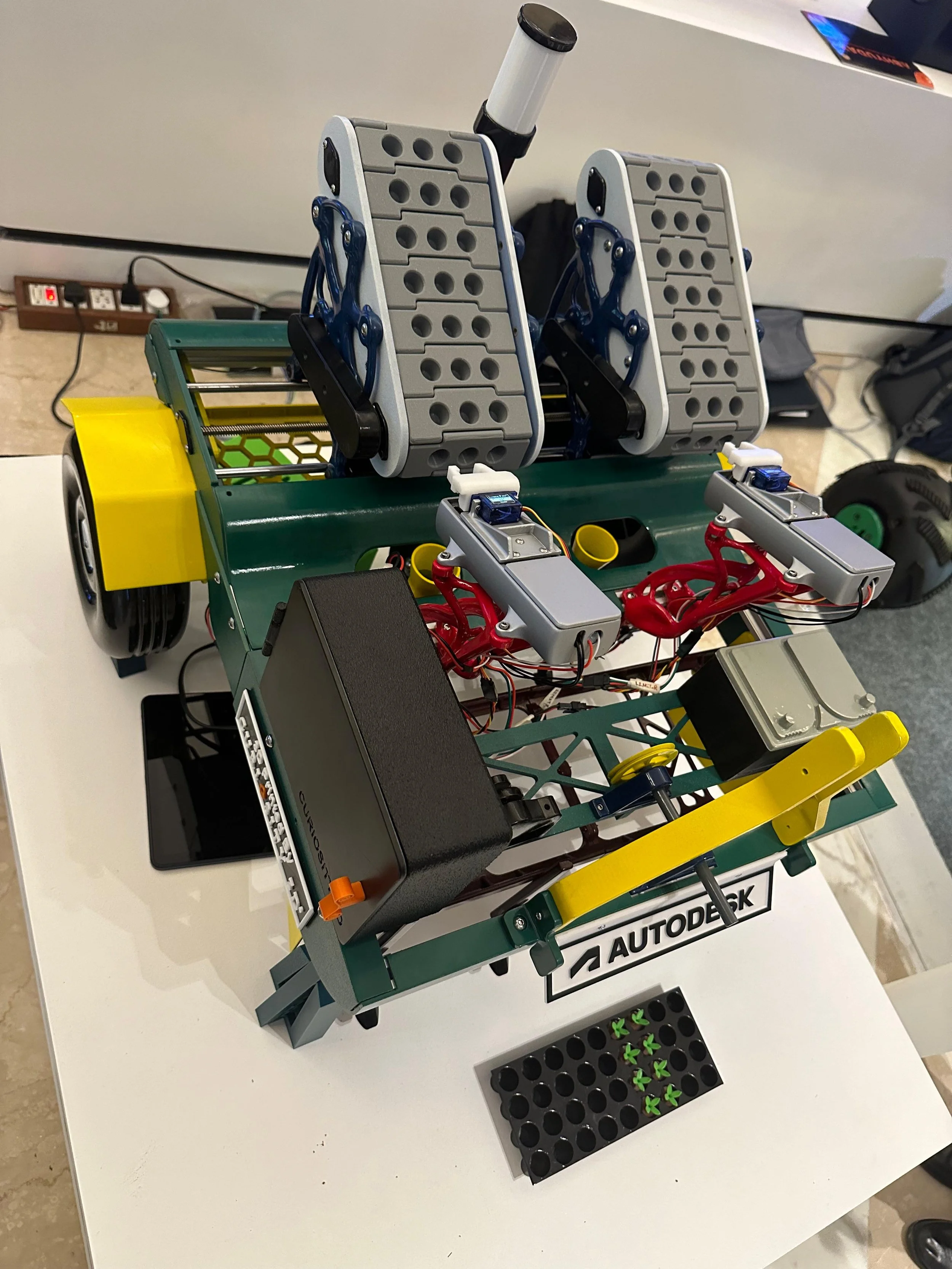 A complex robotic vehicle with metal and plastic parts, electronic components, sensors, and a green base, displayed on a white table with an Autodesk sign. It has two gray foot pedal-like structures on top and multiple wiring connections.