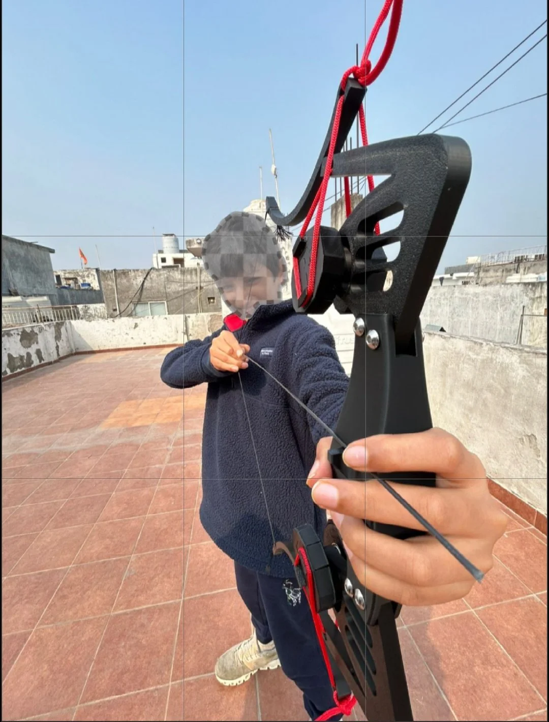 A young boy aiming a black compound bow with red accents on a rooftop. The boy is wearing a blue fleece jacket and in the background, there are buildings and a clear sky.