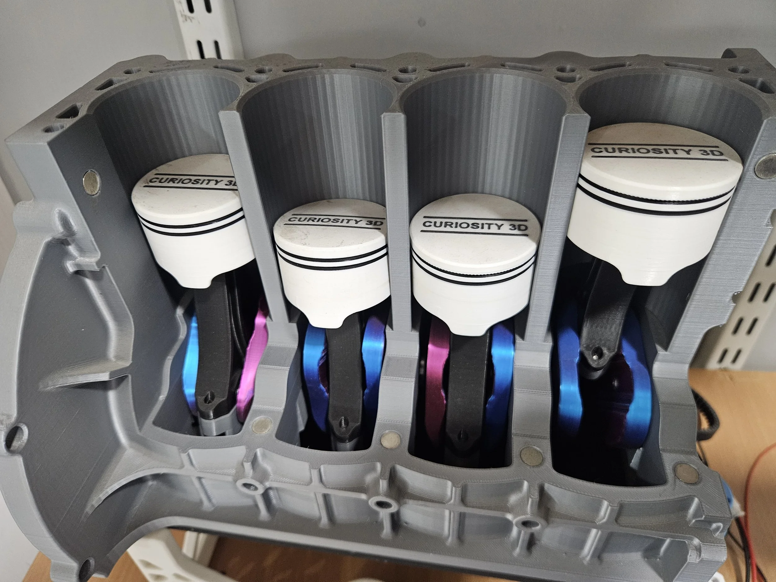 A close-up view of a car engine block with four pistons labeled 'Curiosity 3D' and colorful connecting rods in an aluminum casing.