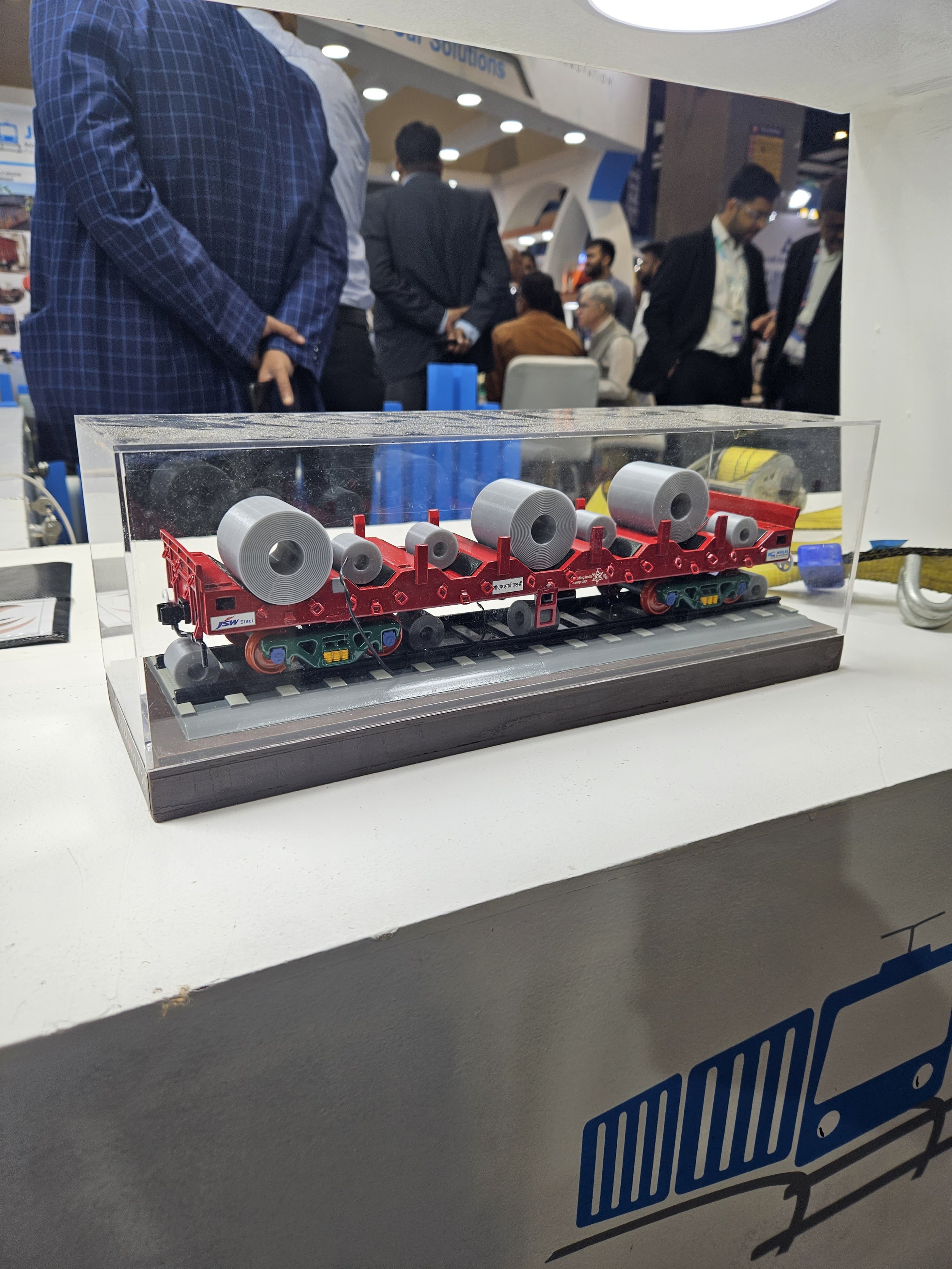 Model train carrying coils displayed in a glass case at an exhibition booth, with people in business attire in the background.