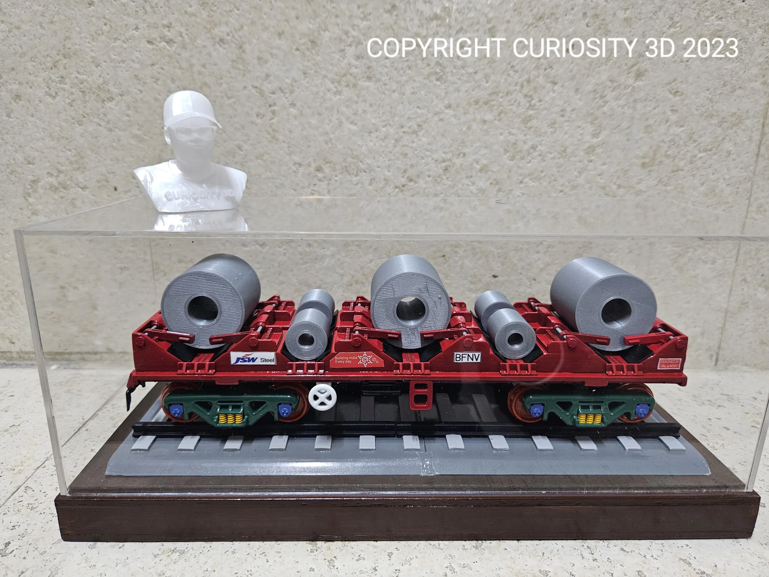 Model train flatbed car carrying steel coils and a sculpture in a display case.