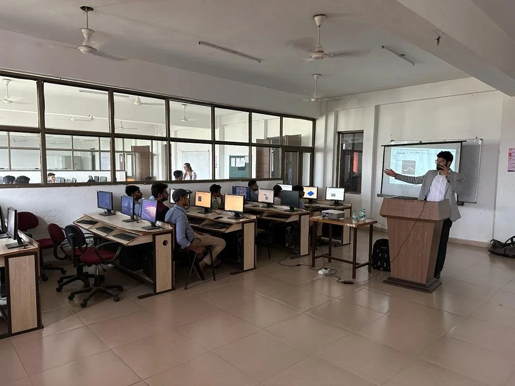 Computer lab with people working on desktops and a person giving a presentation at a podium.