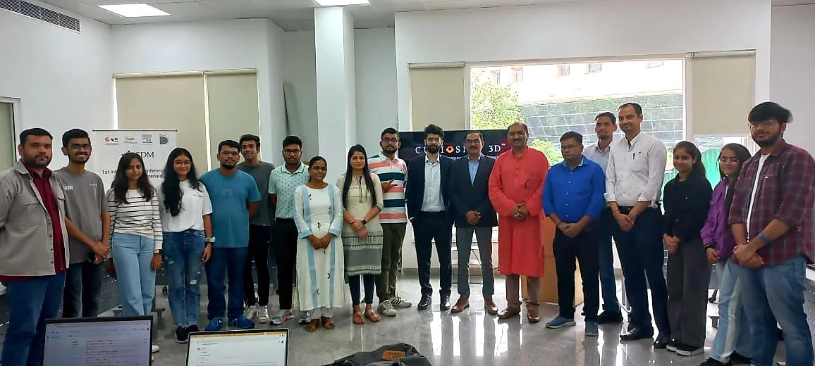 Group of people standing in a large room, smiling for a group photo at an event. Background includes banners and windows. Some are wearing casual clothes, others in formal attire.